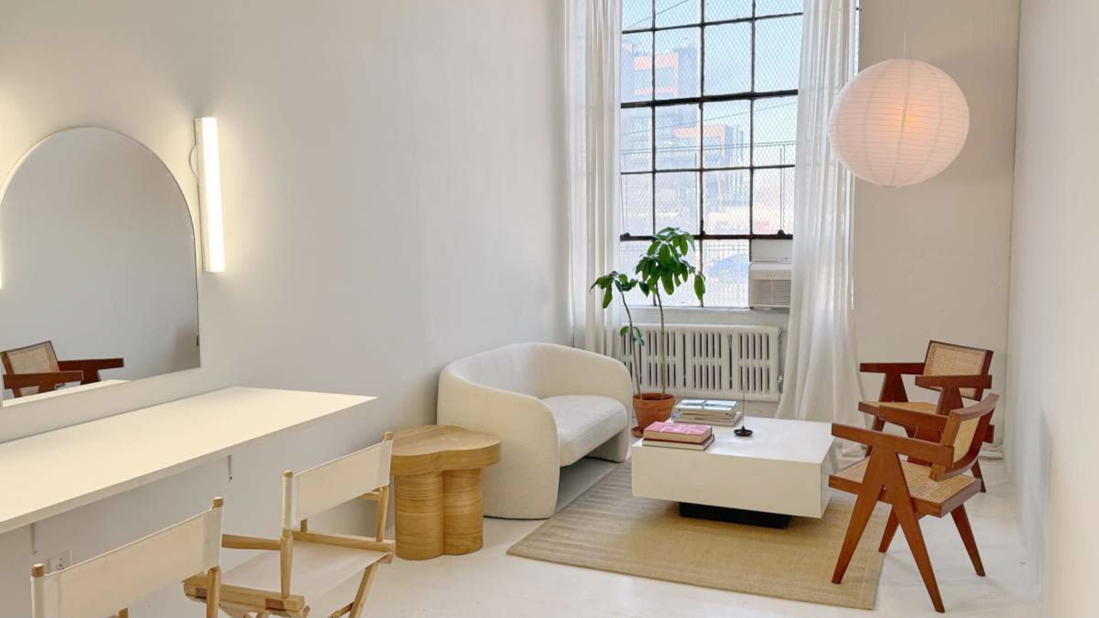 The image shows a minimalist living space featuring a curved sofa, a wooden side table, a small coffee table, and a large window with sheer curtains.