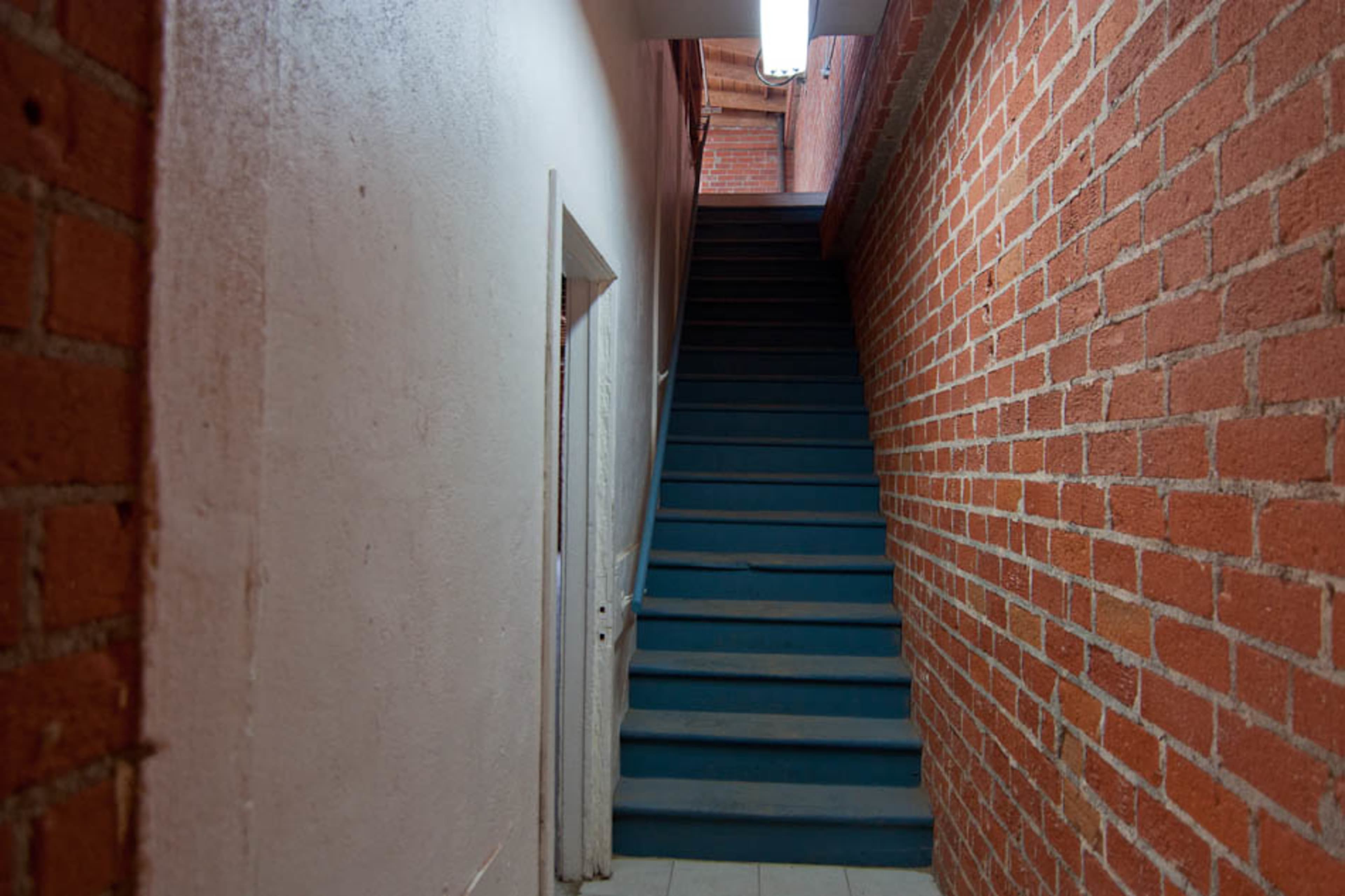A narrow hallway with bare brick walls leads to a staircase at the end, illuminated by a ceiling light.