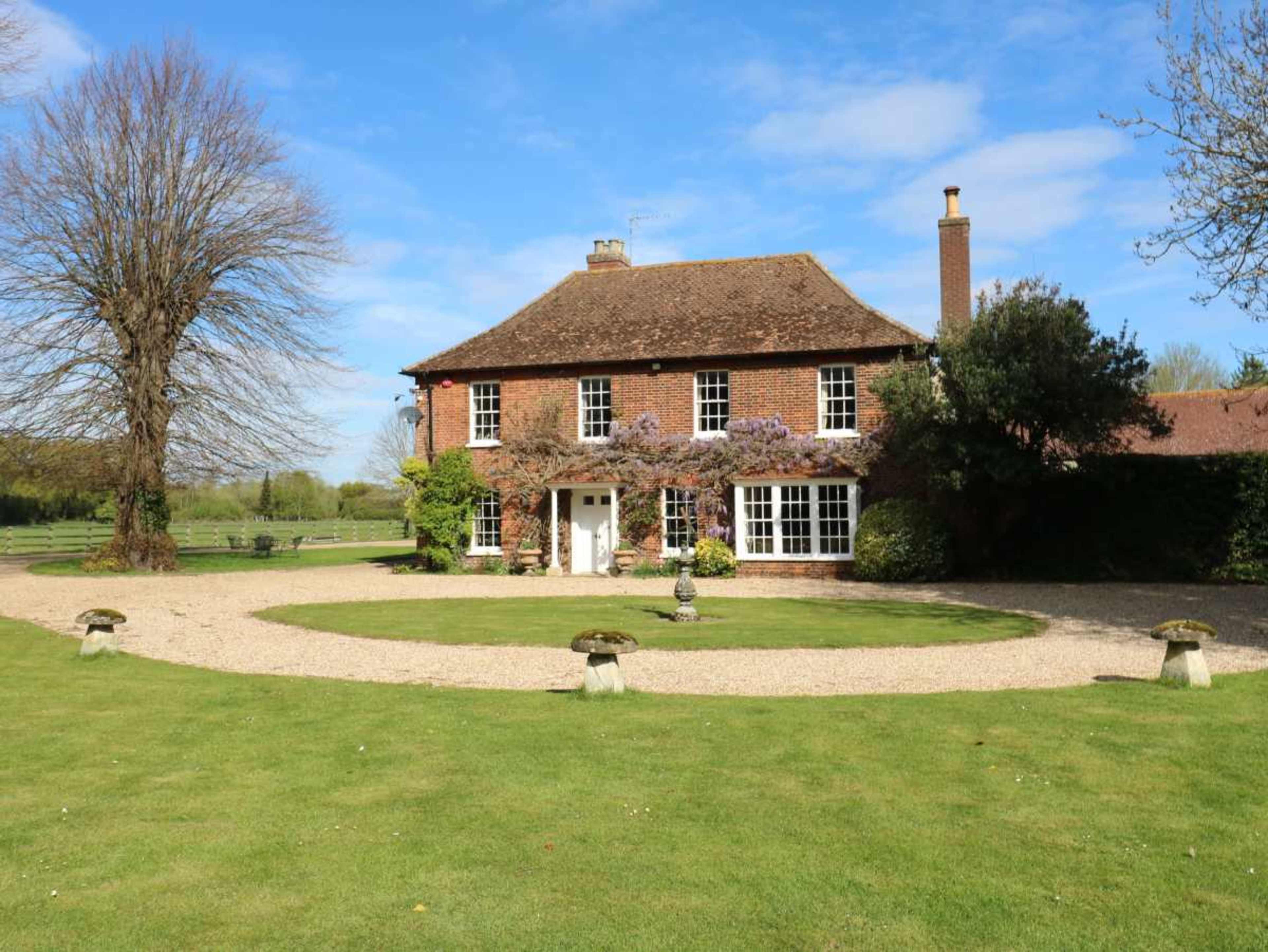The image shows a large brick house with a symmetrical design, surrounded by a circular gravel driveway and well-maintained green lawn, under a clear blue sky.