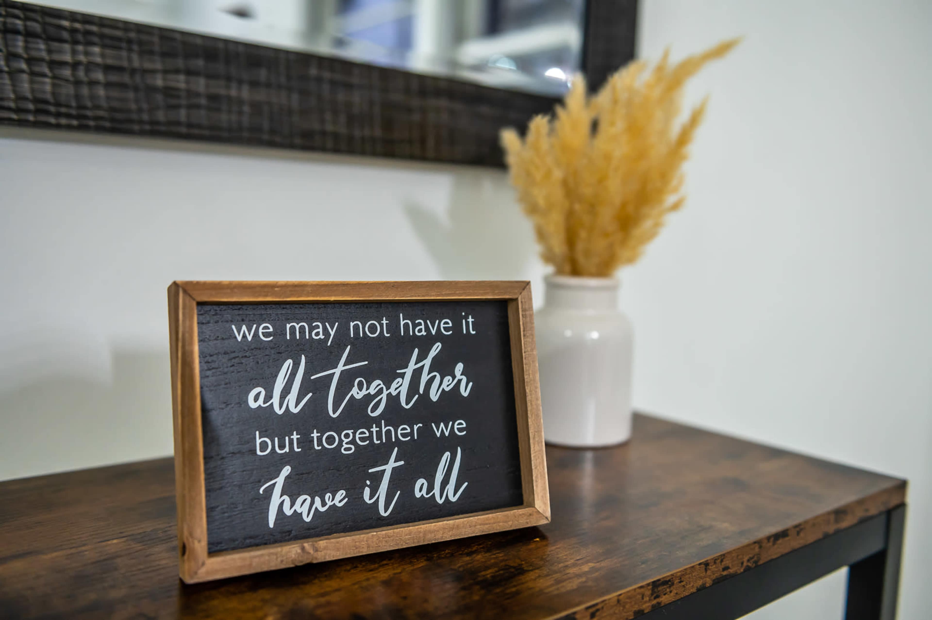 A wooden sign displaying a motivational quote is positioned on a dark wooden table next to a white vase with dried pampas grass, in front of a mirror.