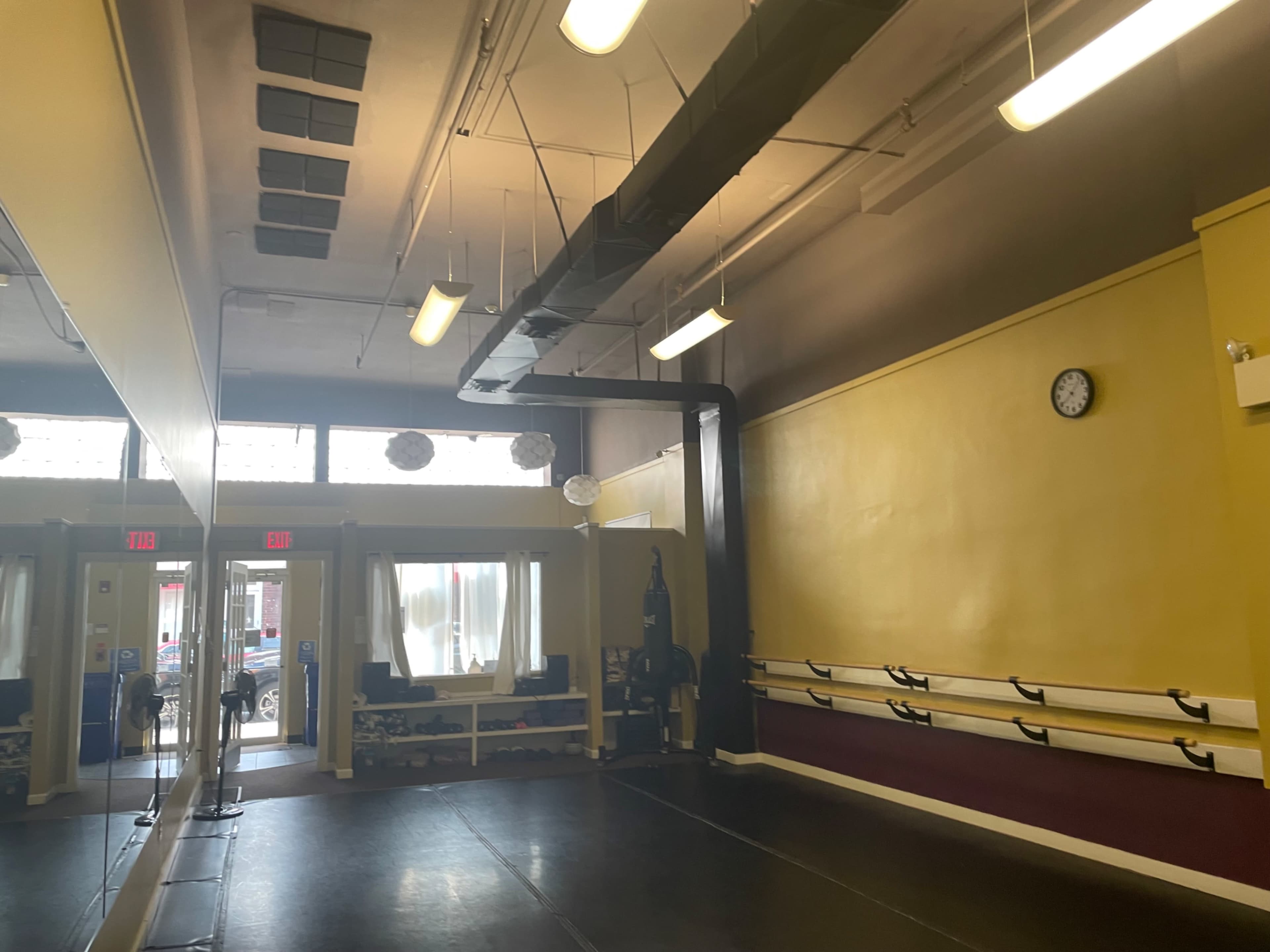 The image depicts a well-lit dance studio featuring mirrored walls, a wooden barre along one side, and a clock mounted on the yellow wall.