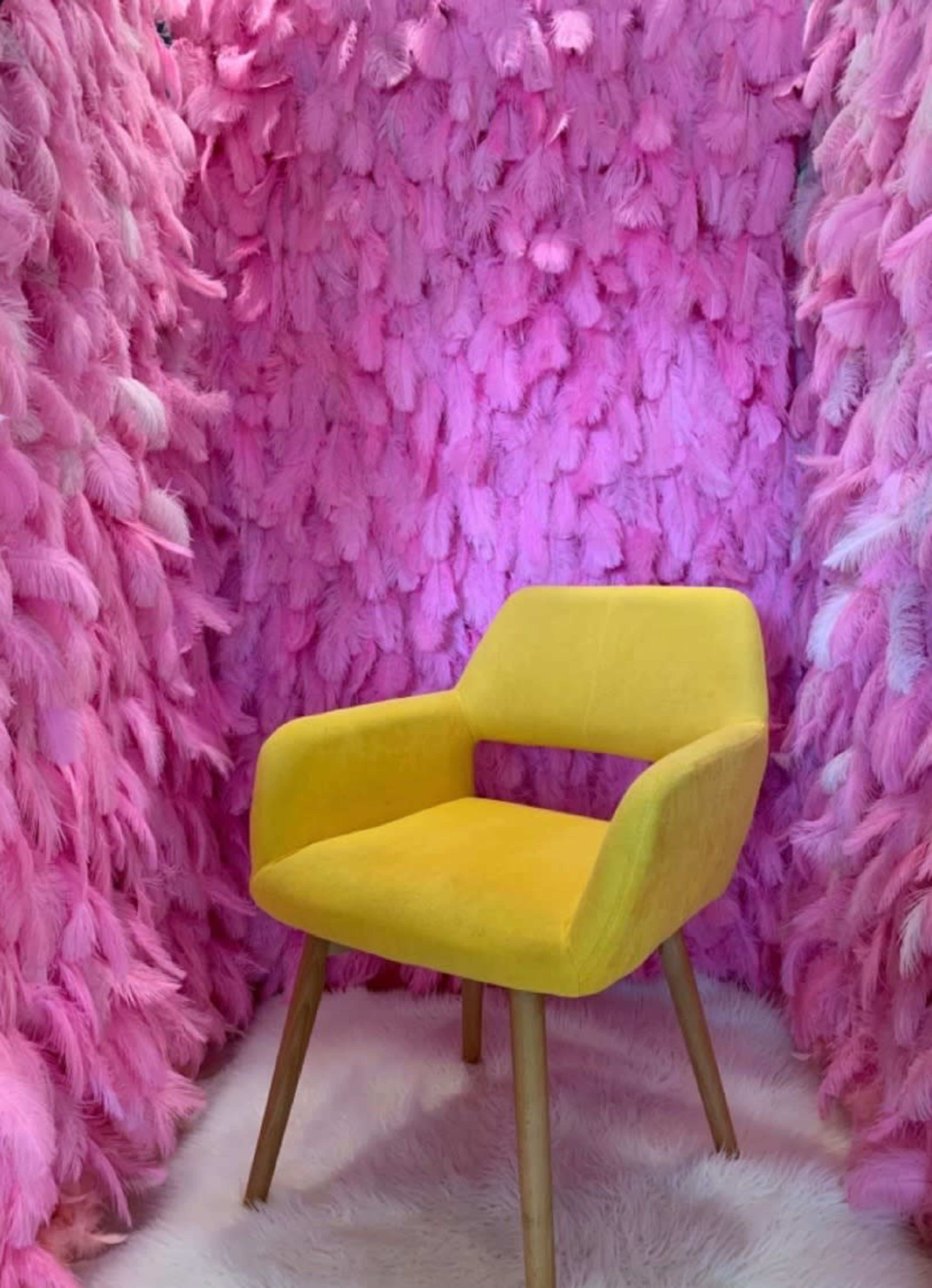 A yellow chair is positioned in front of a wall made of pink feathers.