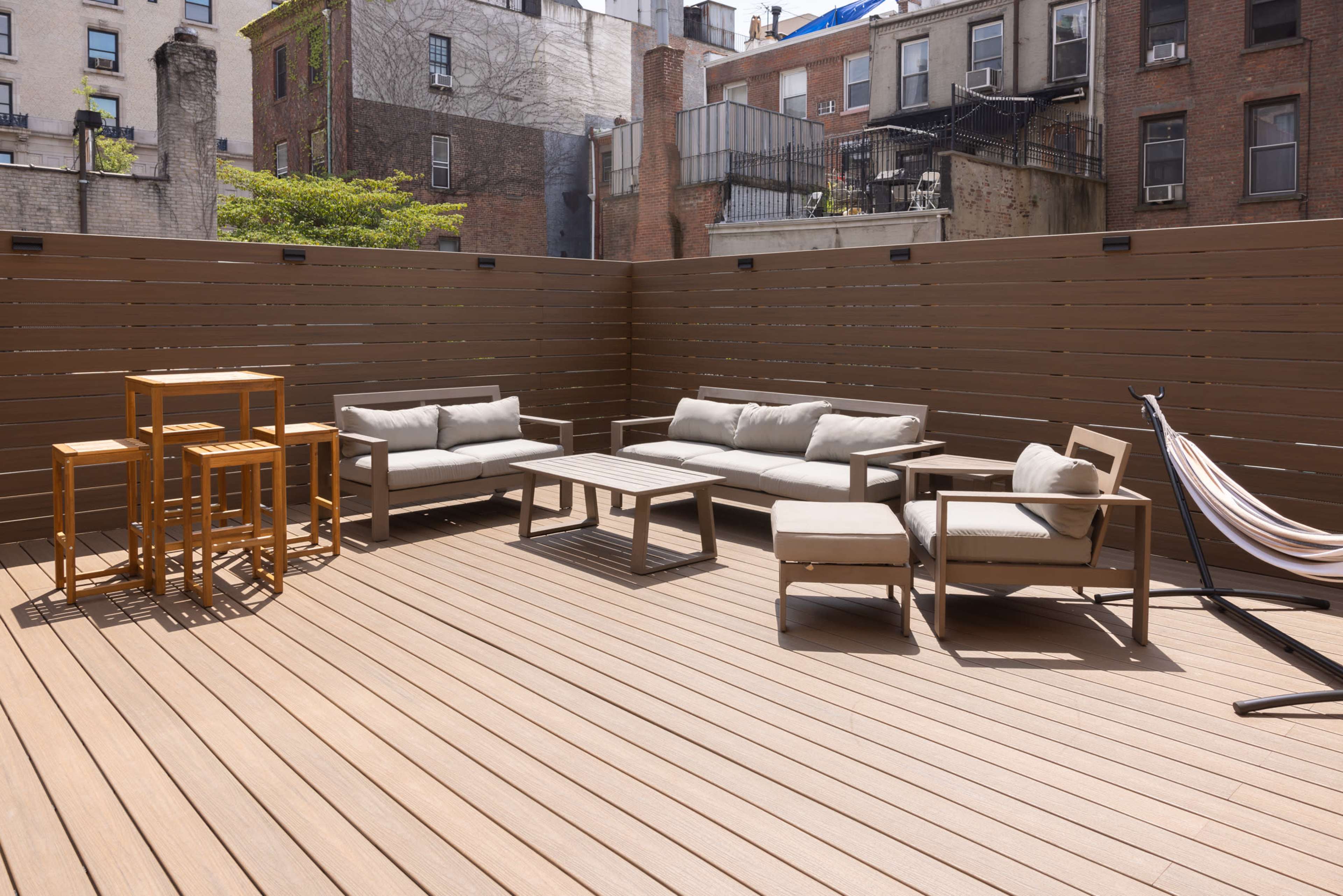 The image shows a modern outdoor patio area with wooden decking, featuring a seating arrangement of a sofa, armchair, and coffee table, along with a hammock.