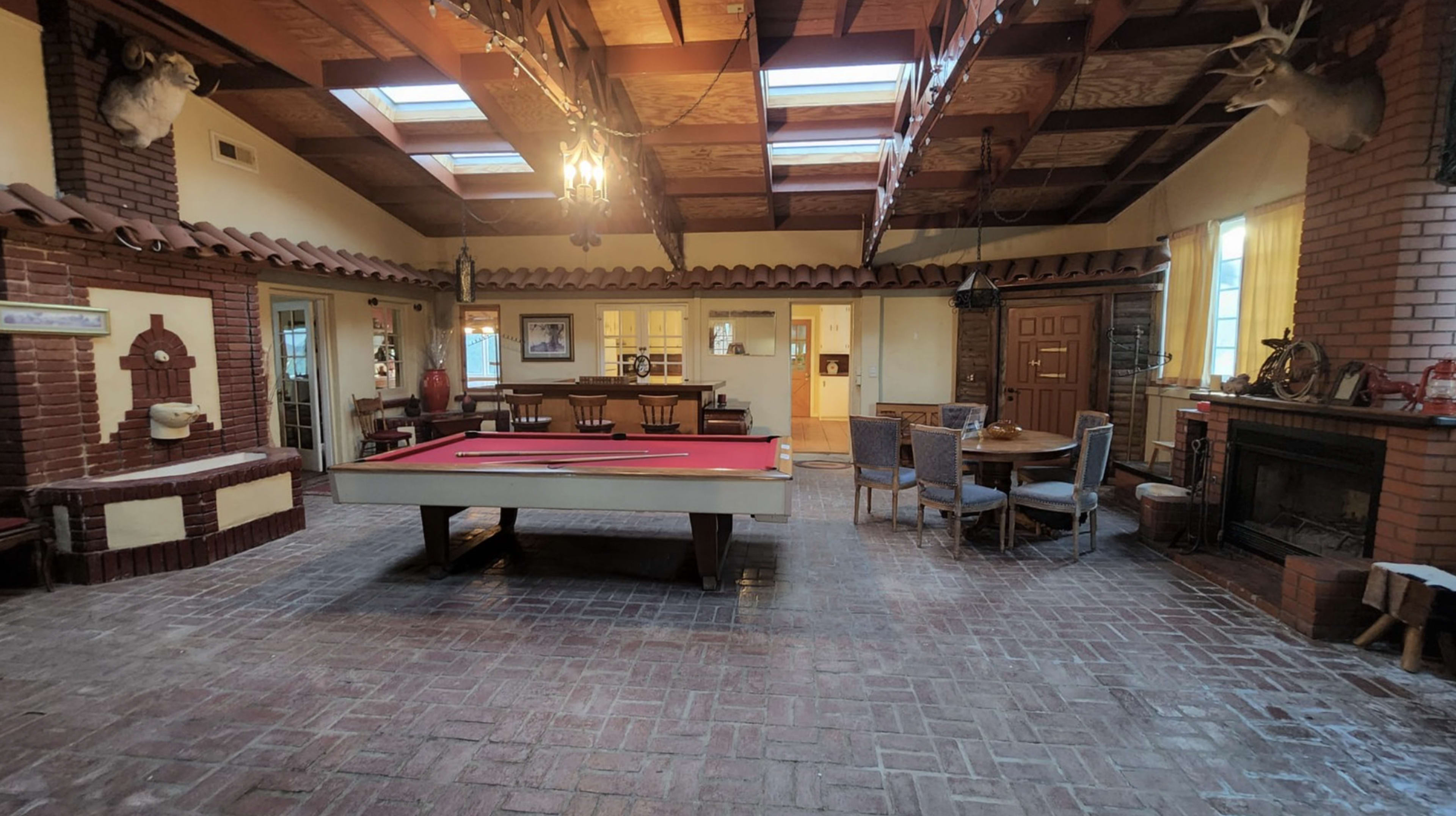 A spacious room featuring a red pool table, brick flooring, wooden beams overhead, and a dining area with a circular table and chairs.