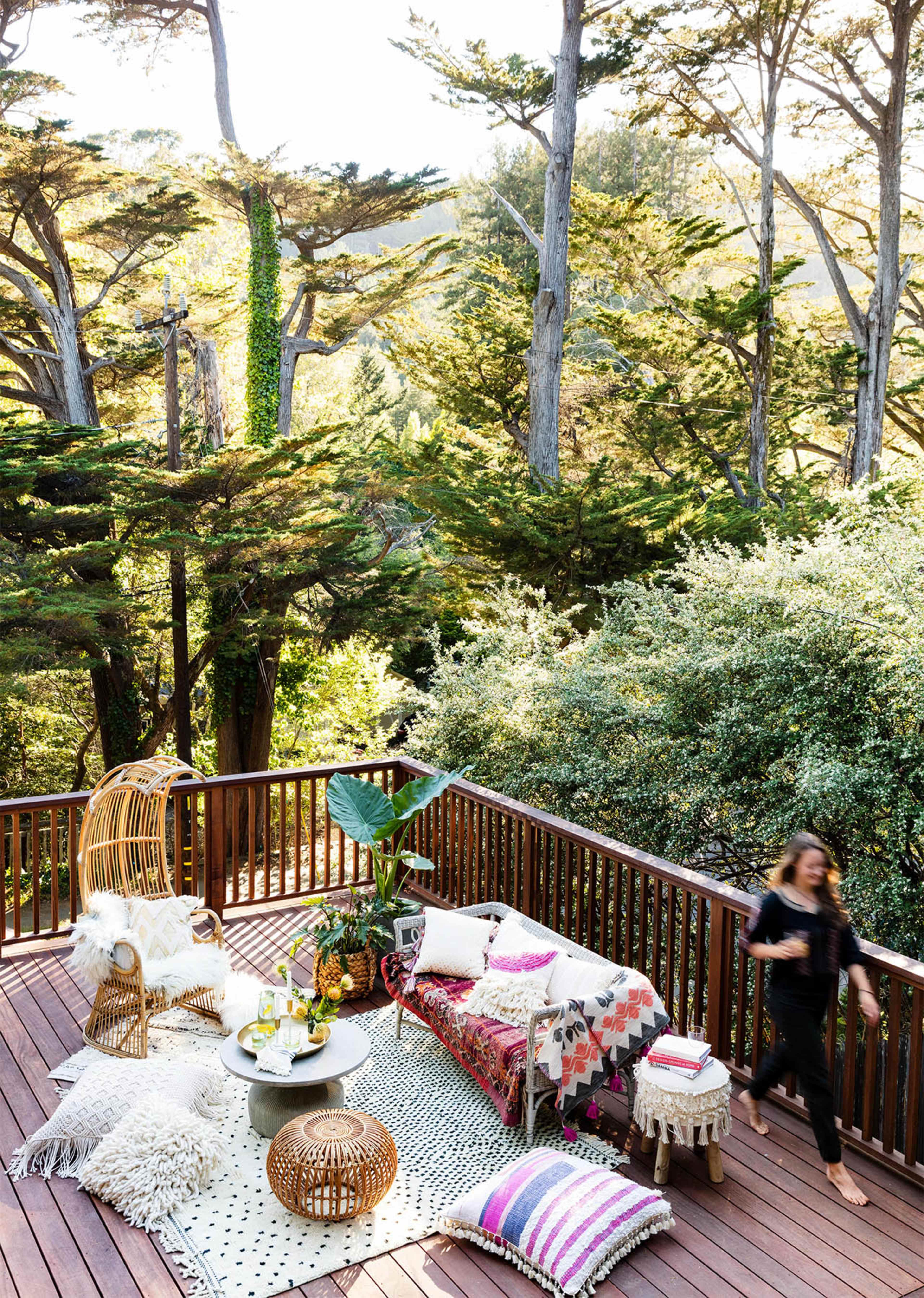 A wooden deck features a cozy seating area with decorative pillows, a table holding drinks, and surrounded by lush greenery and tall trees.