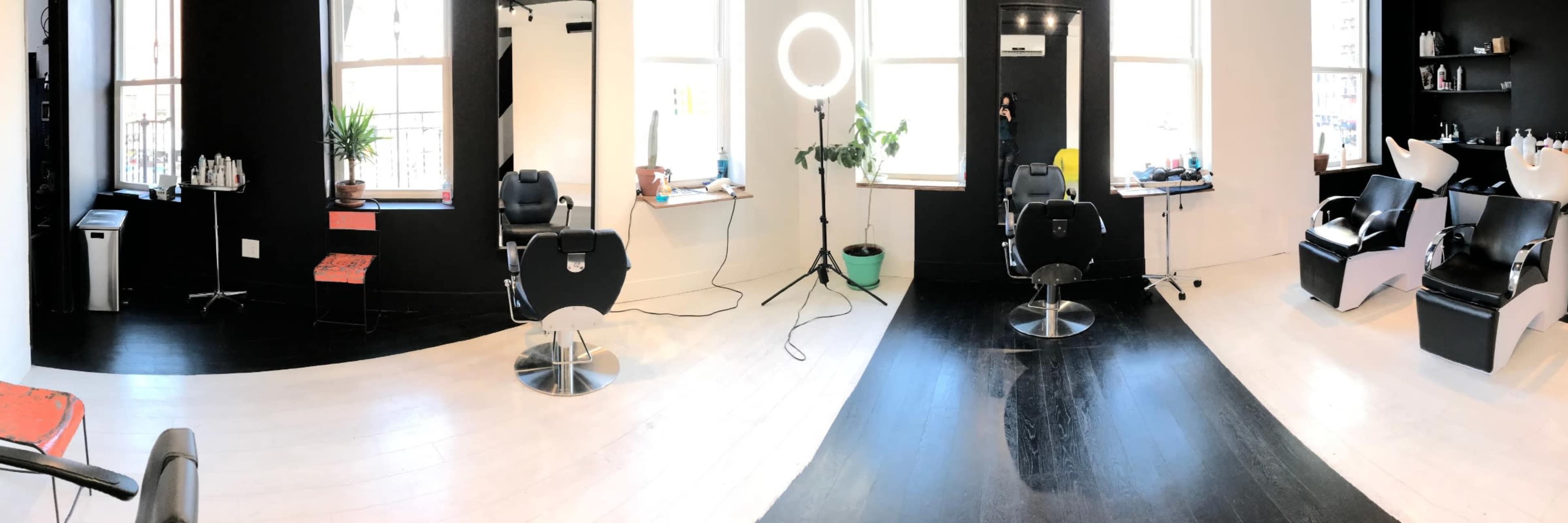 The image shows a modern hair salon interior with black and white flooring, two styling chairs, and a large ring light near a window.