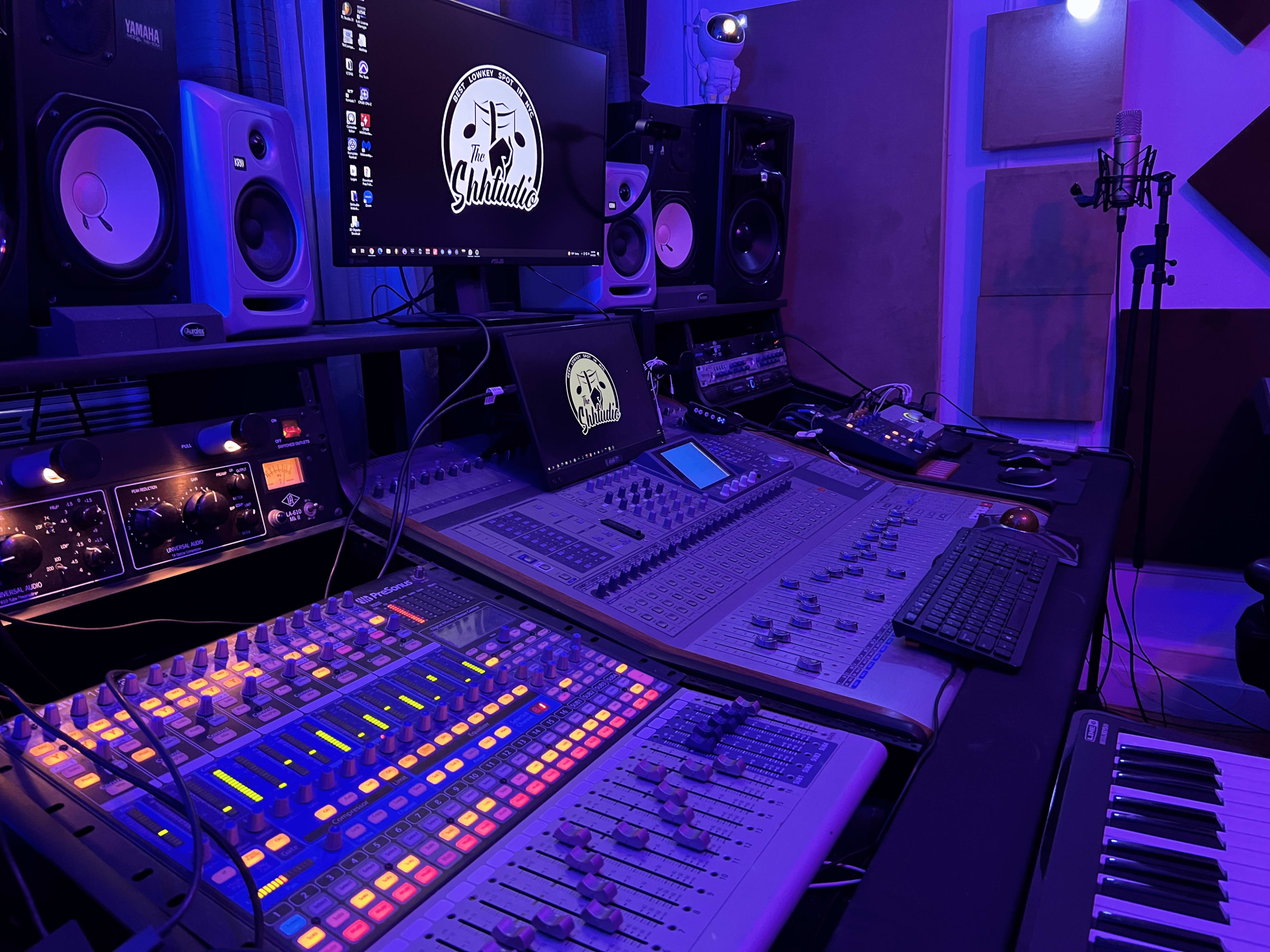A music production studio with a mixing console, computer monitor, and various audio equipment surrounded by blue lighting.