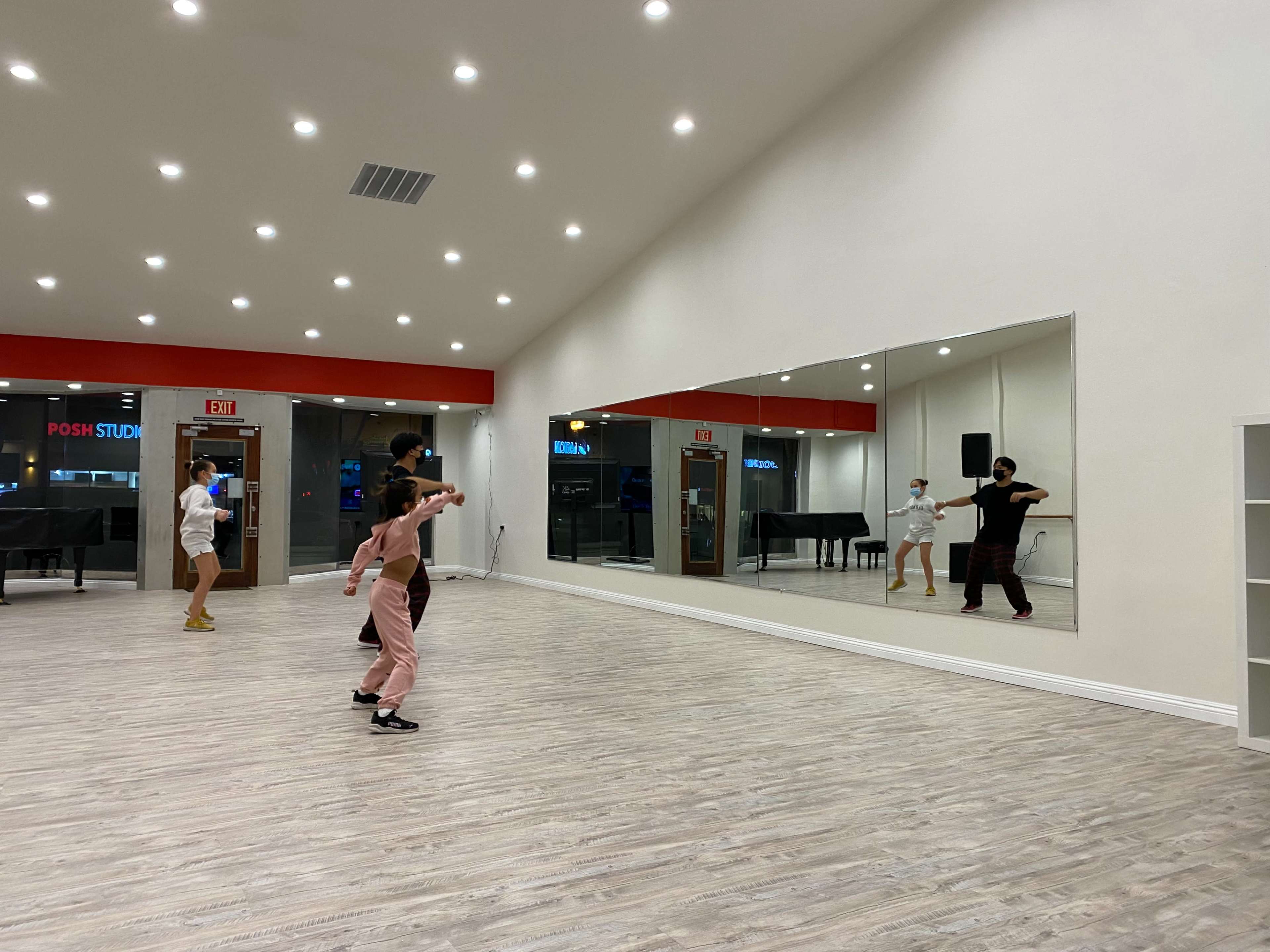 A group of dancers practices in a brightly lit studio with a large mirror and wooden flooring.