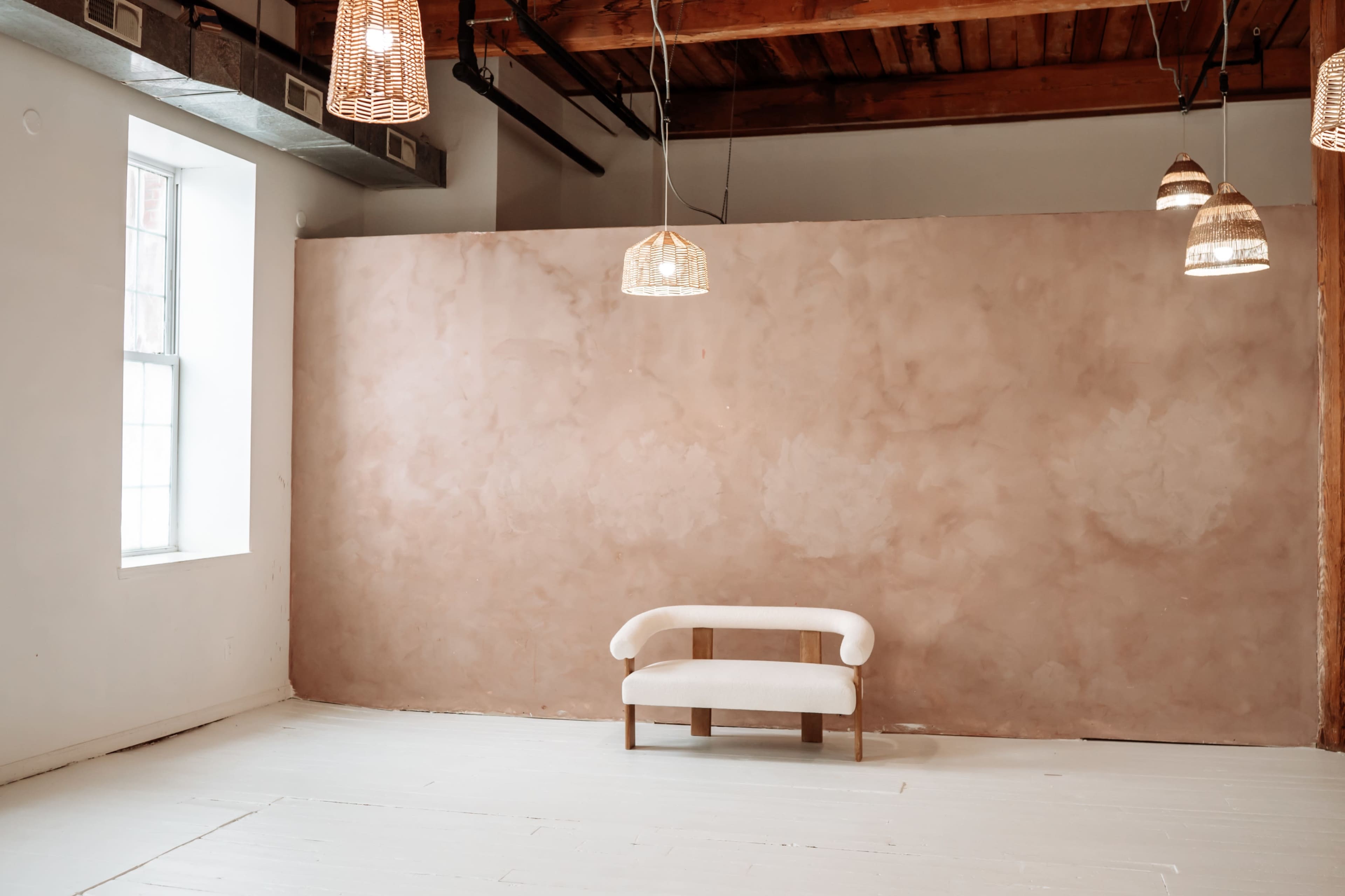 The image shows a minimalist interior space featuring a simple white sofa in front of a textured light pink wall, illuminated by hanging pendant lights.