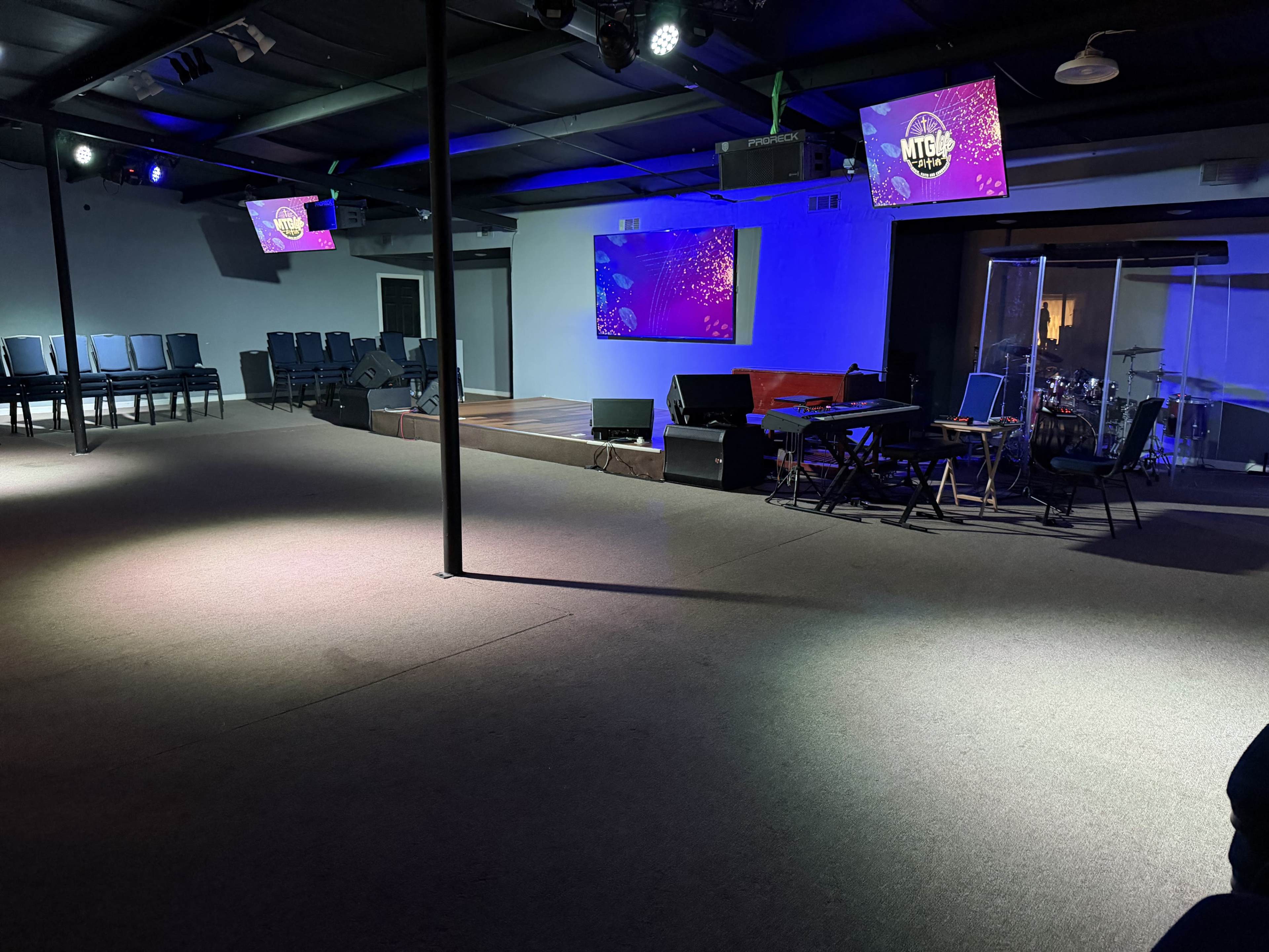 The image shows an empty indoor space with a stage, seating area, and a band setup, illuminated by colored lights.