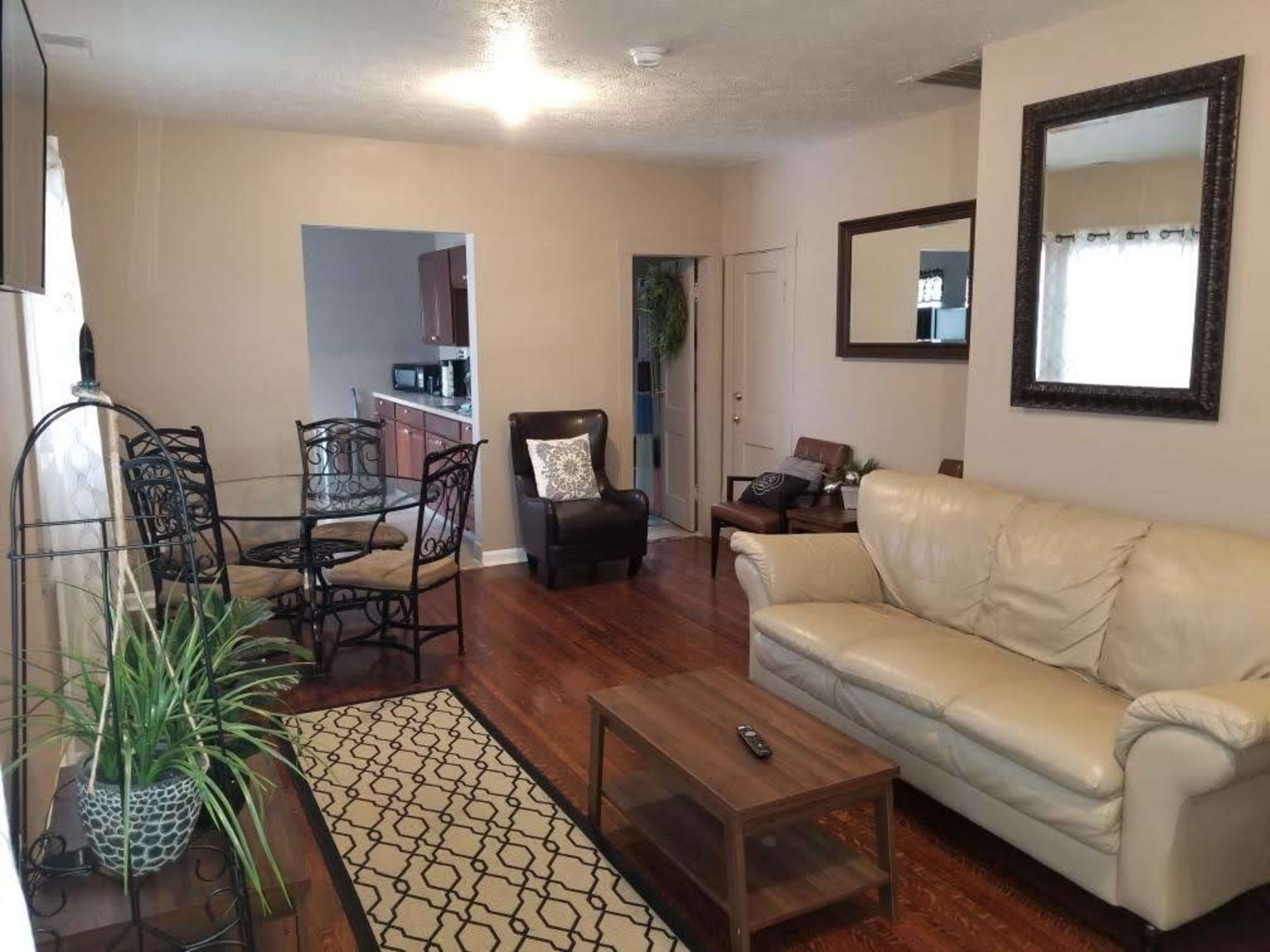 A living room with a leather sofa, a wooden coffee table, a dining area with a glass-top table and chairs, a large mirror on the wall, and a doorway leading to a kitchen.