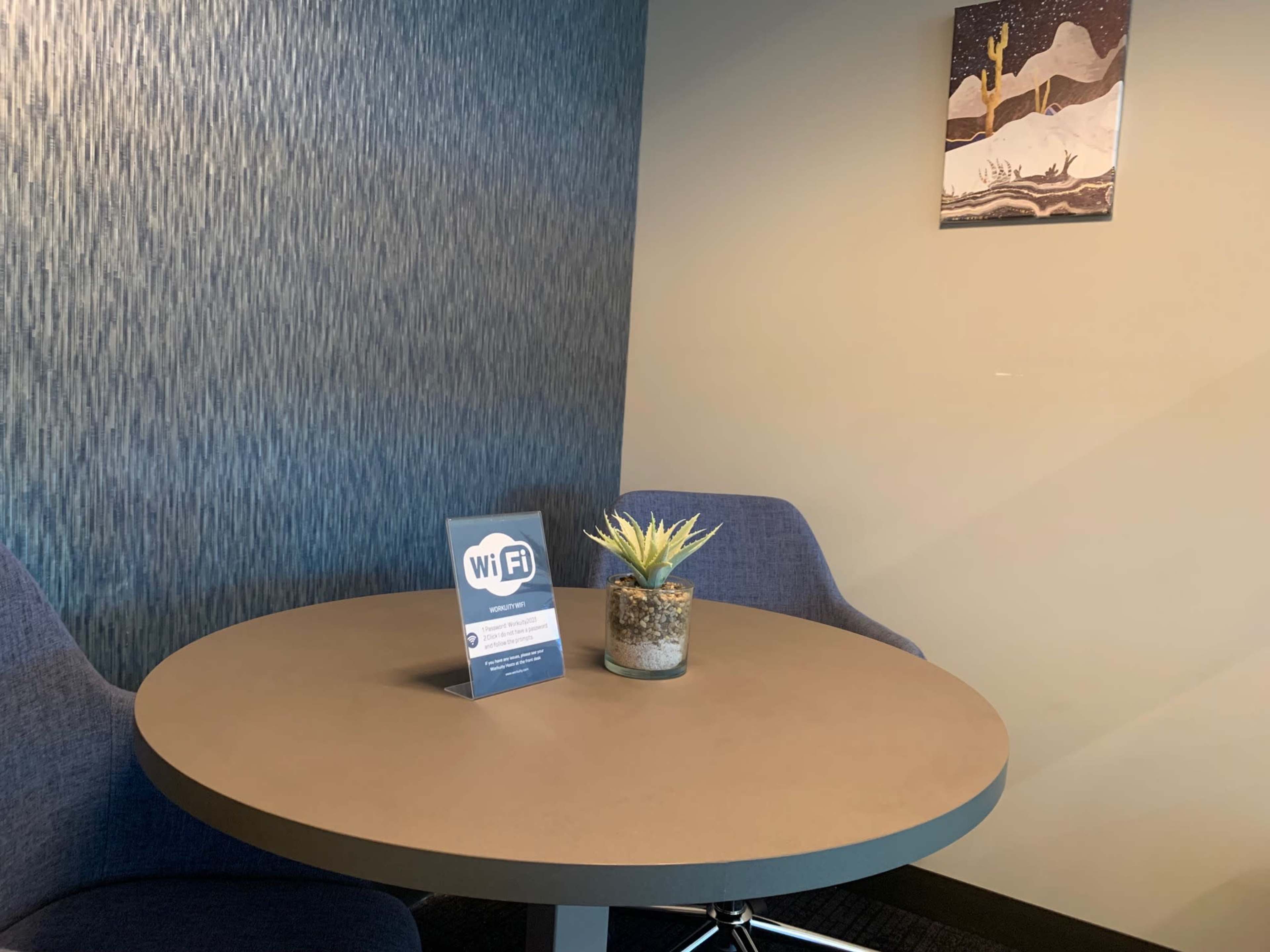 A small table with two chairs features a Wi-Fi sign and a potted plant, set against a textured wall and a piece of art depicting a desert scene.
