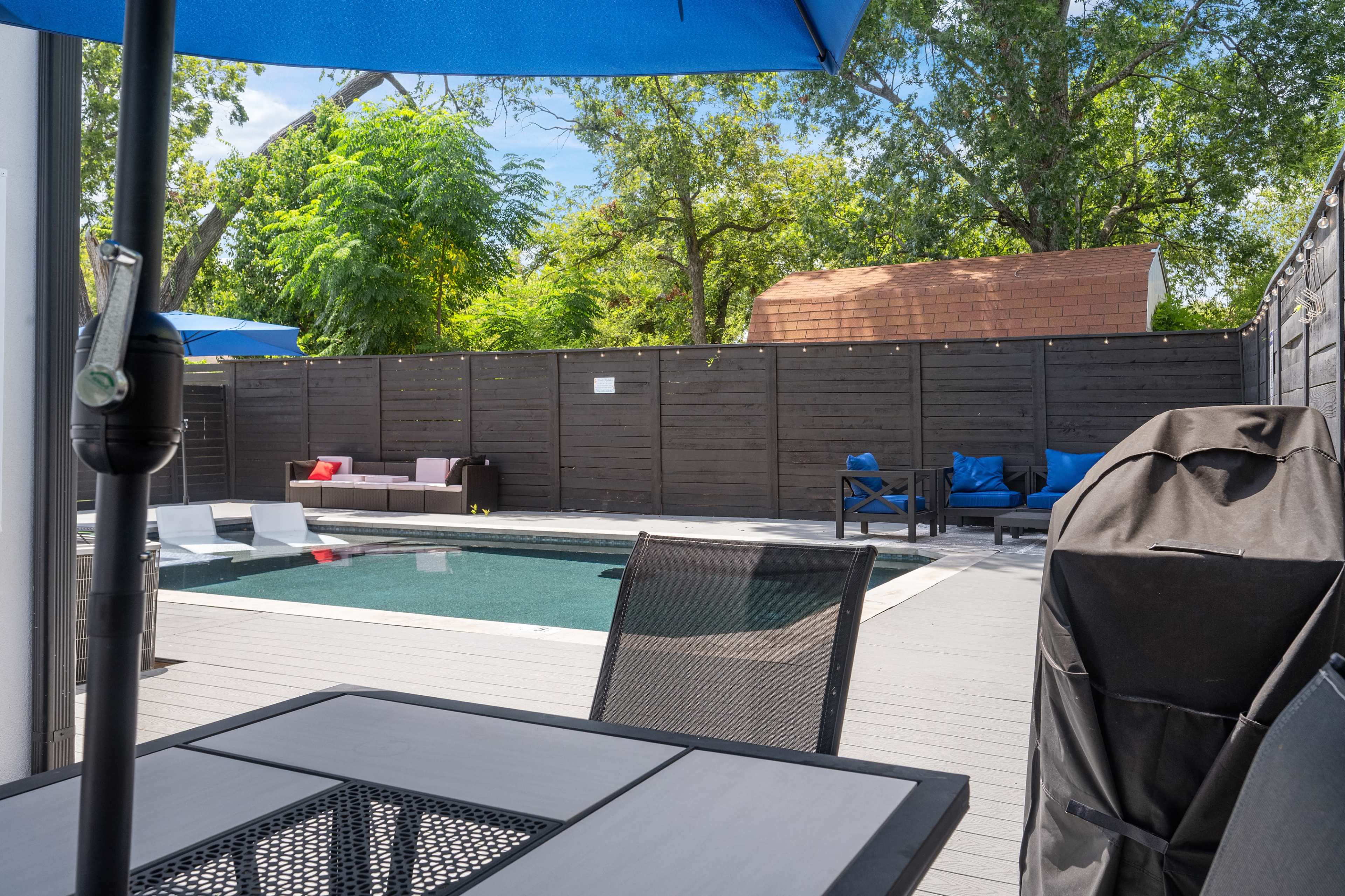 The image depicts a backyard pool area with a swimming pool surrounded by lounge chairs and an outdoor dining table, all enclosed by a wooden fence and shaded by large umbrellas.