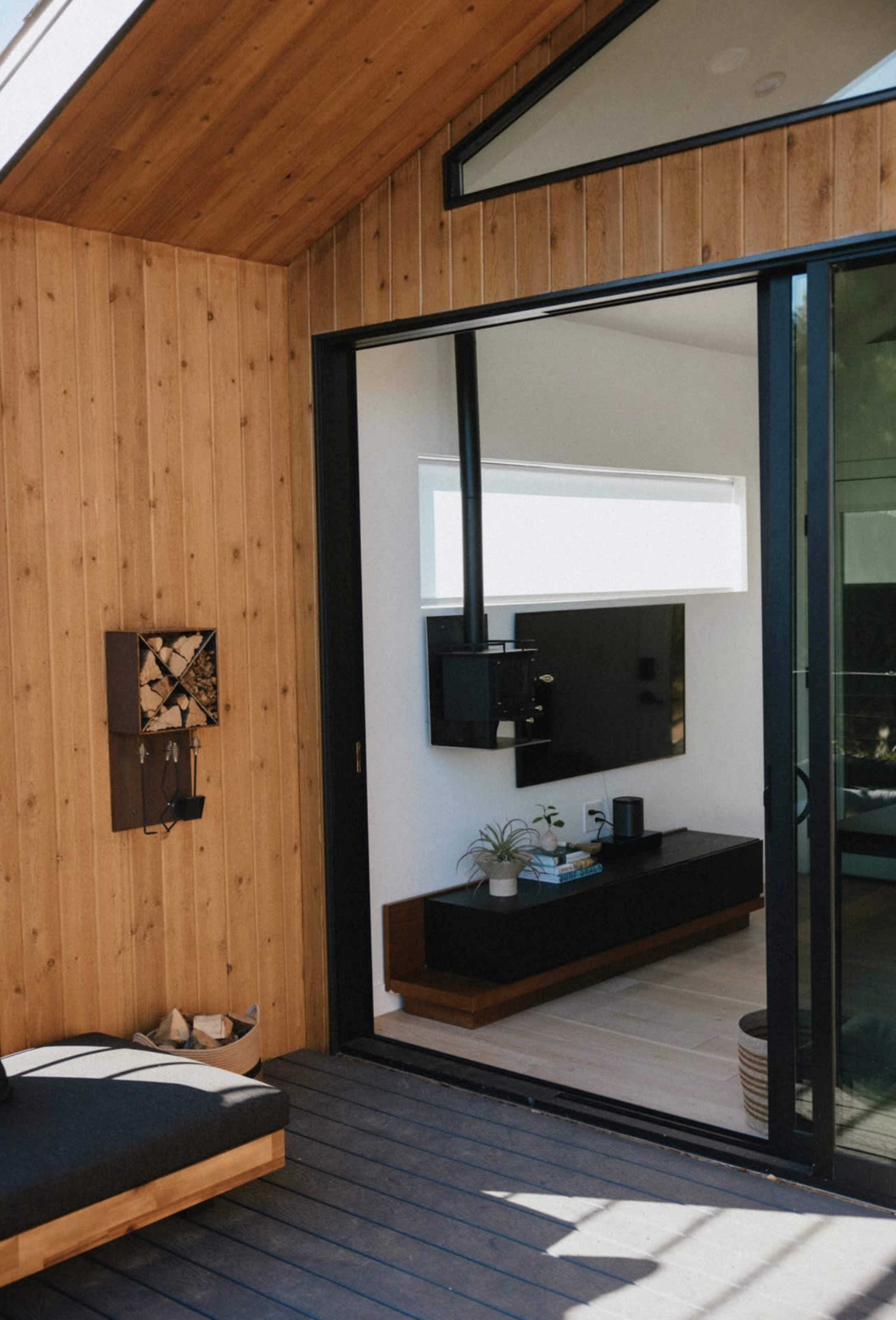 A wooden exterior wall with large glass doors that open to a modern living space featuring a black entertainment unit and indoor plants.