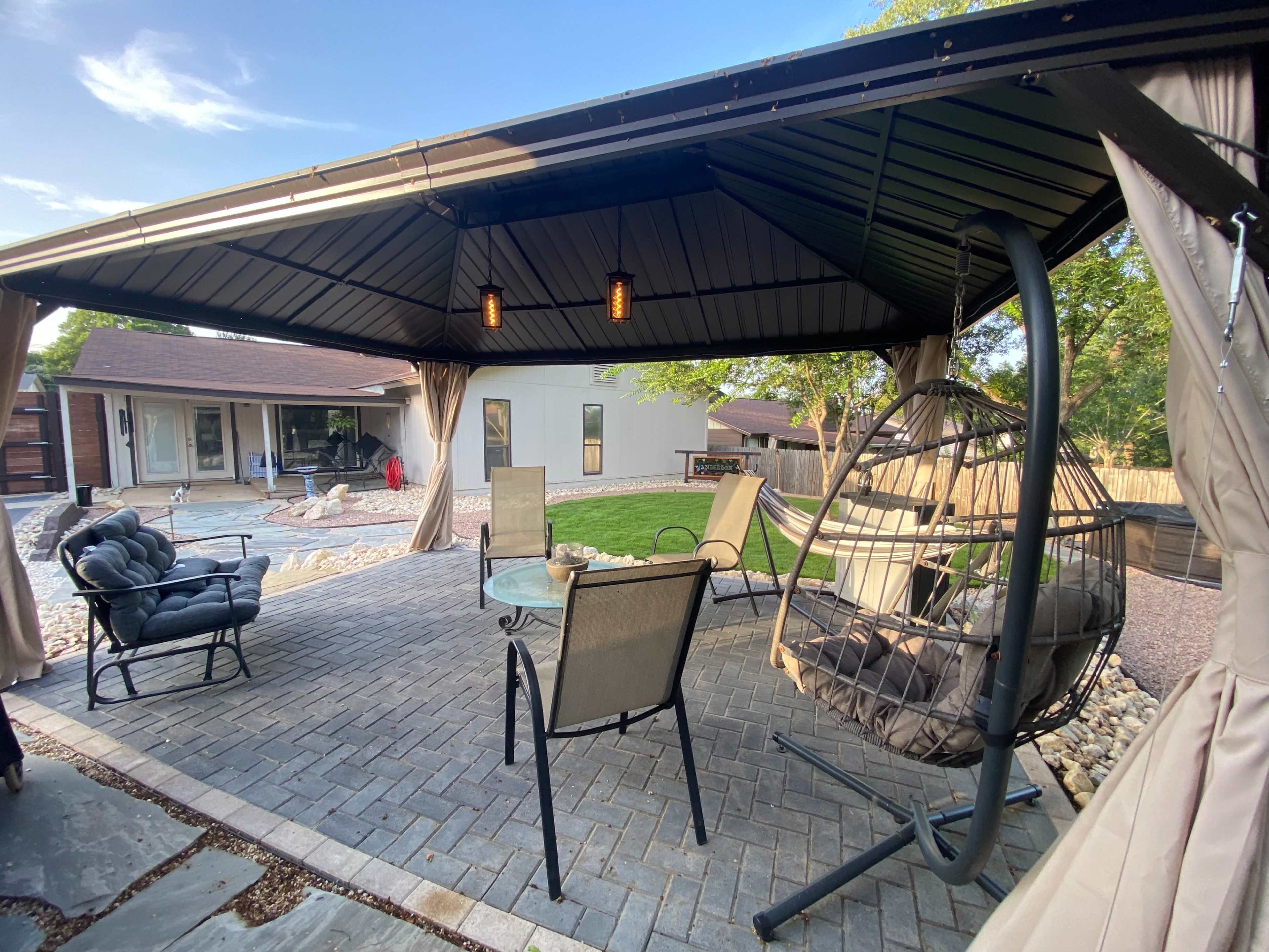 The image shows a gazebo with seating arrangements and a hanging chair, overlooking a landscaped backyard.