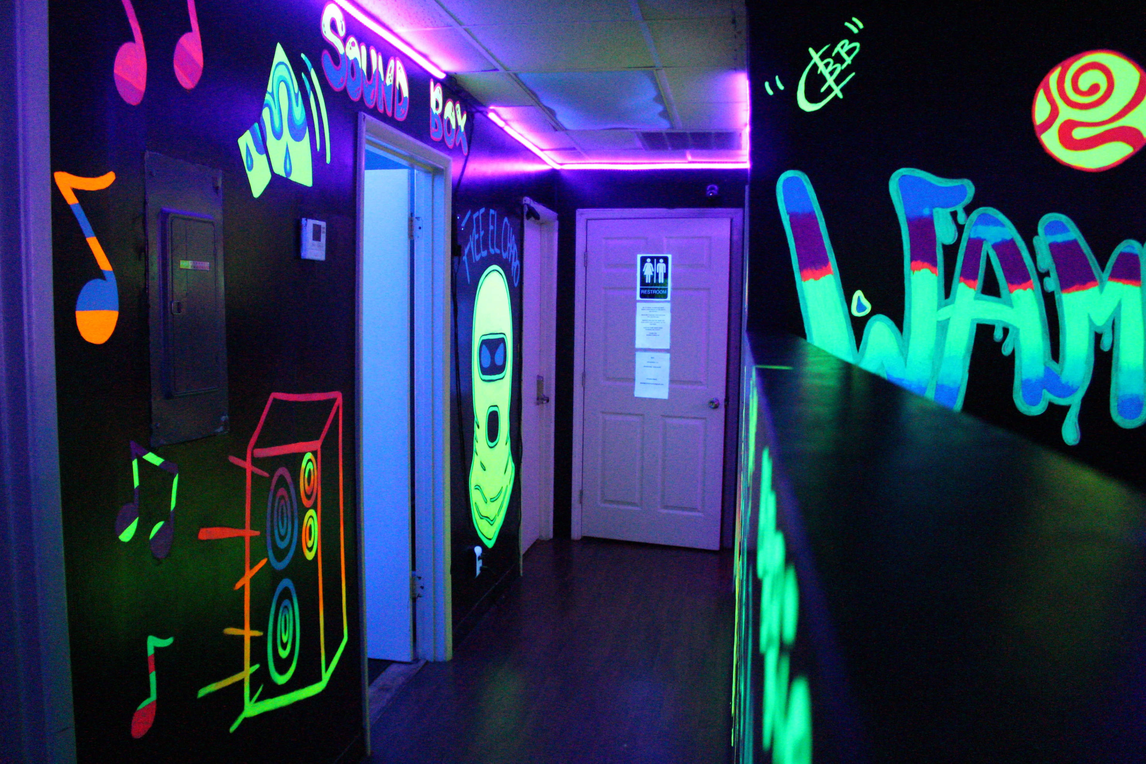 A dimly lit hallway features neon graffiti, including music notes and a cartoonish figure, with purple fluorescent lights illuminating the space.