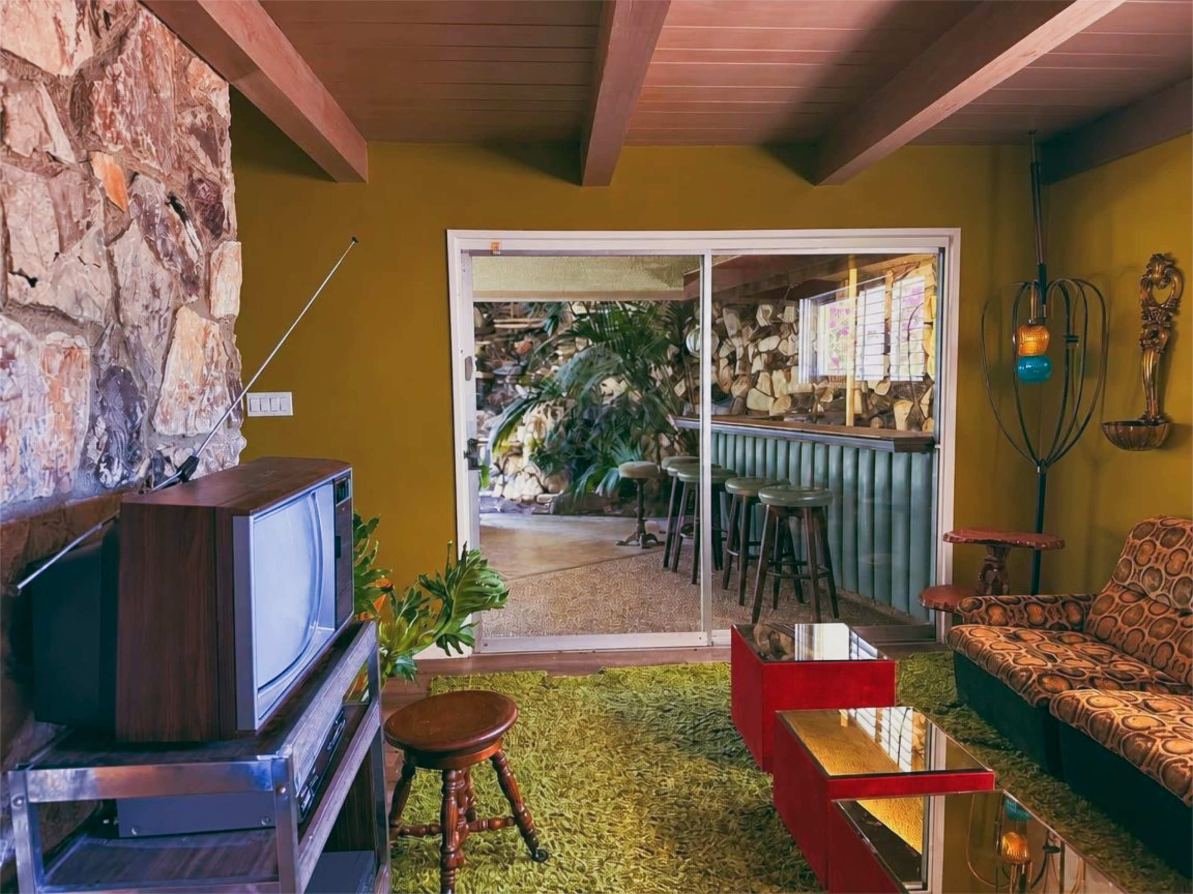 The image shows a living room with a vintage television, a green shag rug, a mid-century modern couch, and an open sliding glass door leading to an outdoor area with a bar.