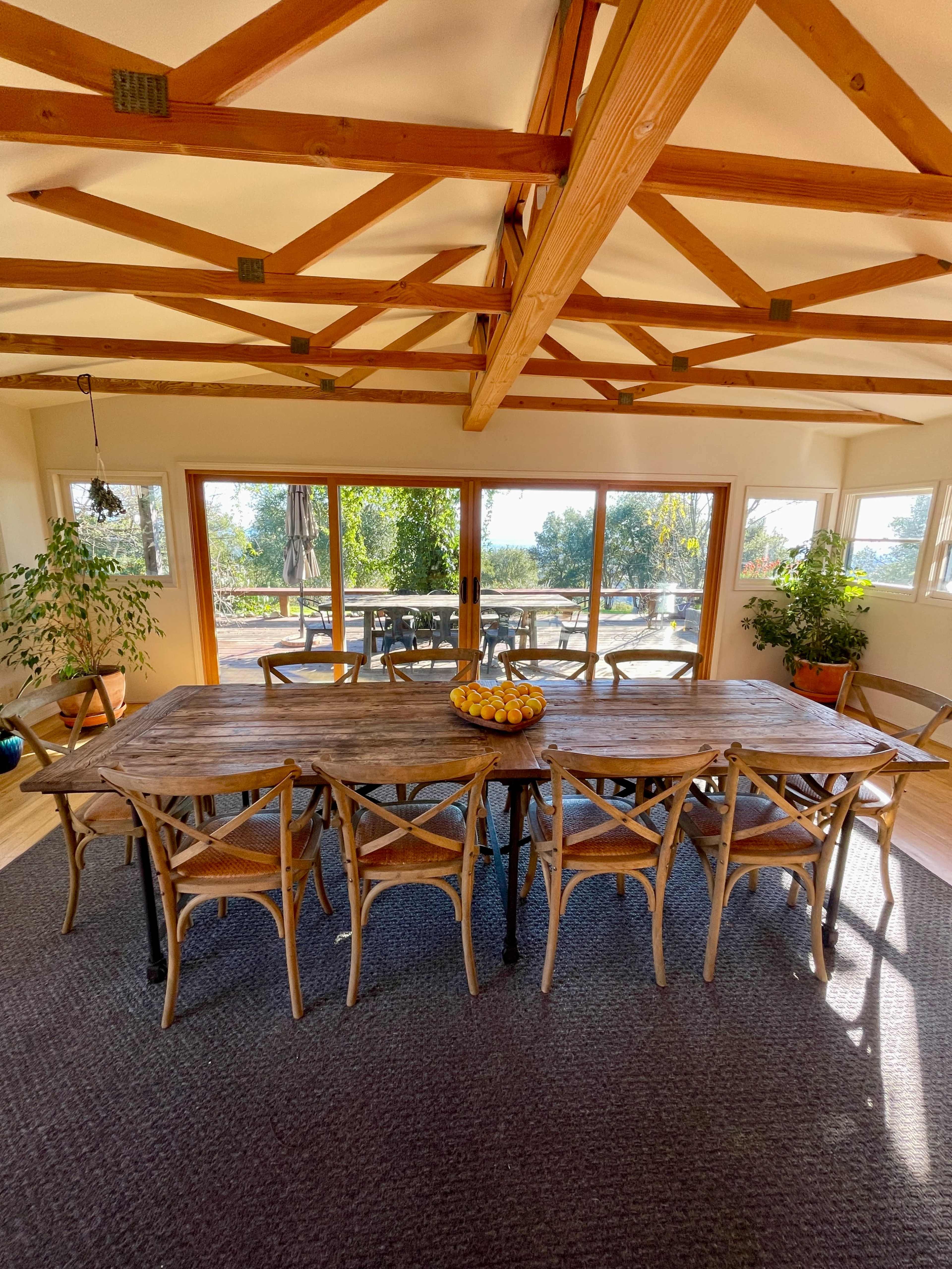 A spacious dining area features a large wooden table surrounded by chairs, with large windows overlooking an outdoor terrace and greenery.