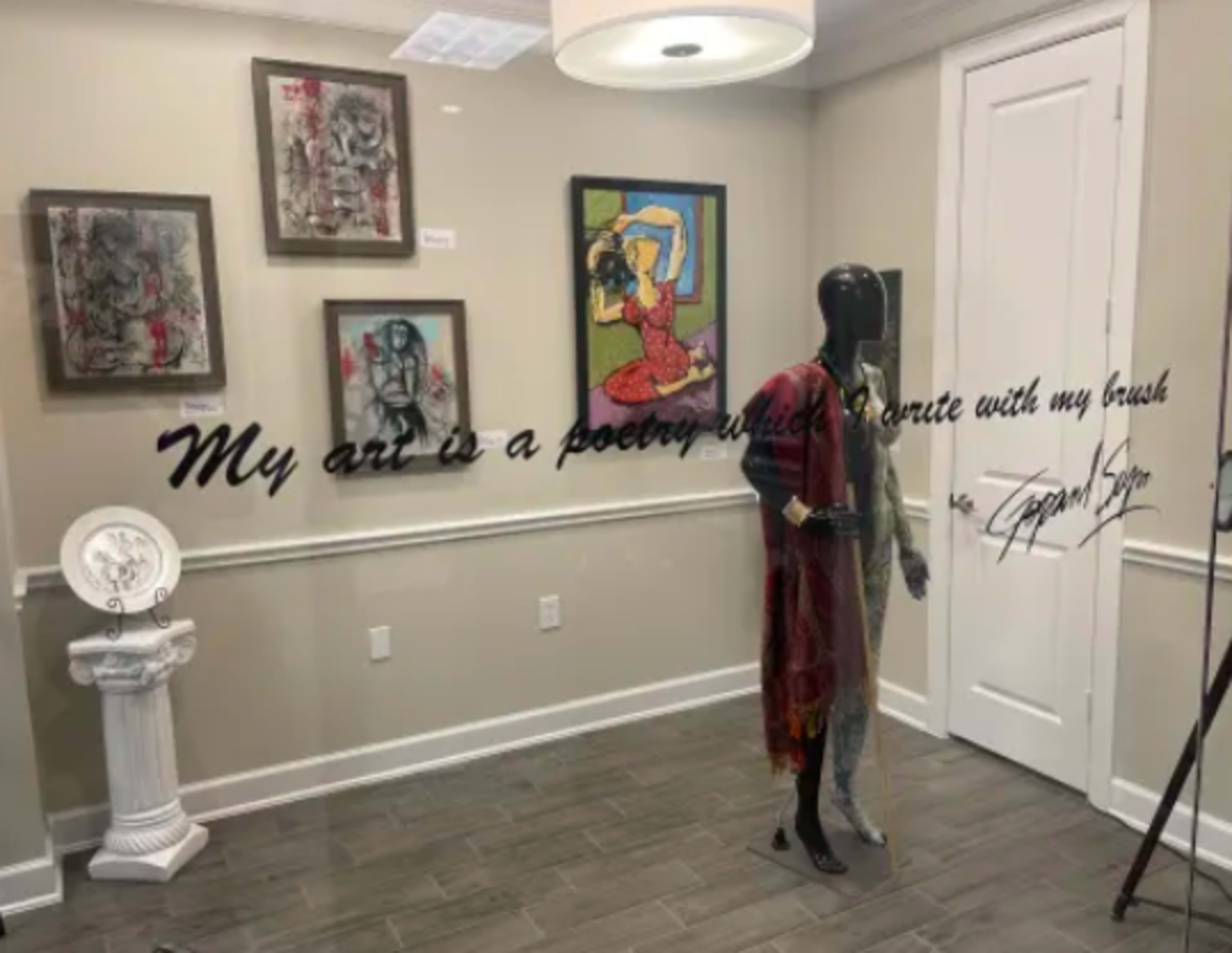 The image features a gallery space with framed artworks on the wall, a mannequin dressed in clothing, and a decorative quote displayed prominently.