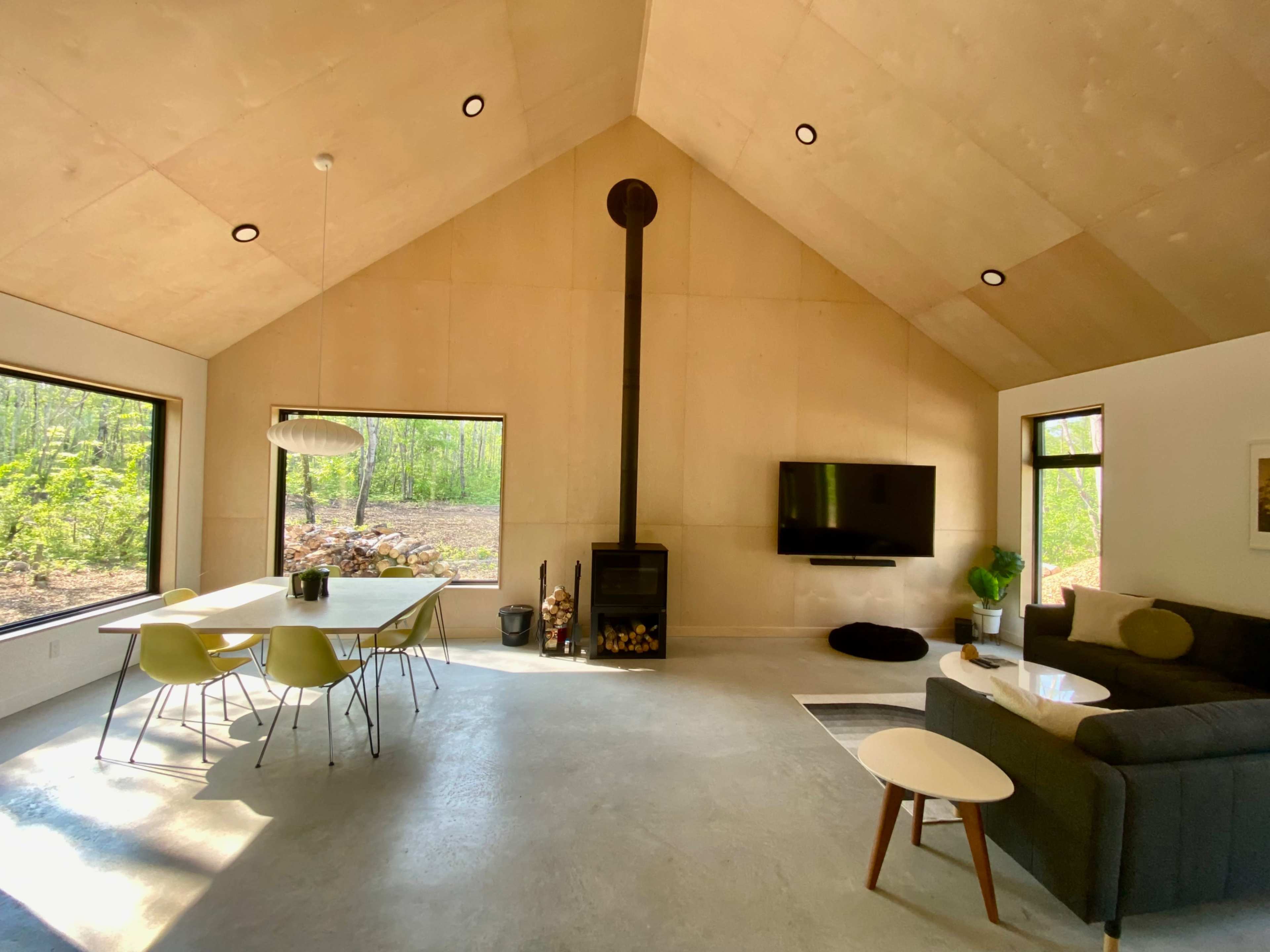 A modern interior living space featuring a dining area with a table and chairs, a sectional sofa, a wall-mounted TV, and a wood-burning stove against a backdrop of large windows overlooking a wooded area.