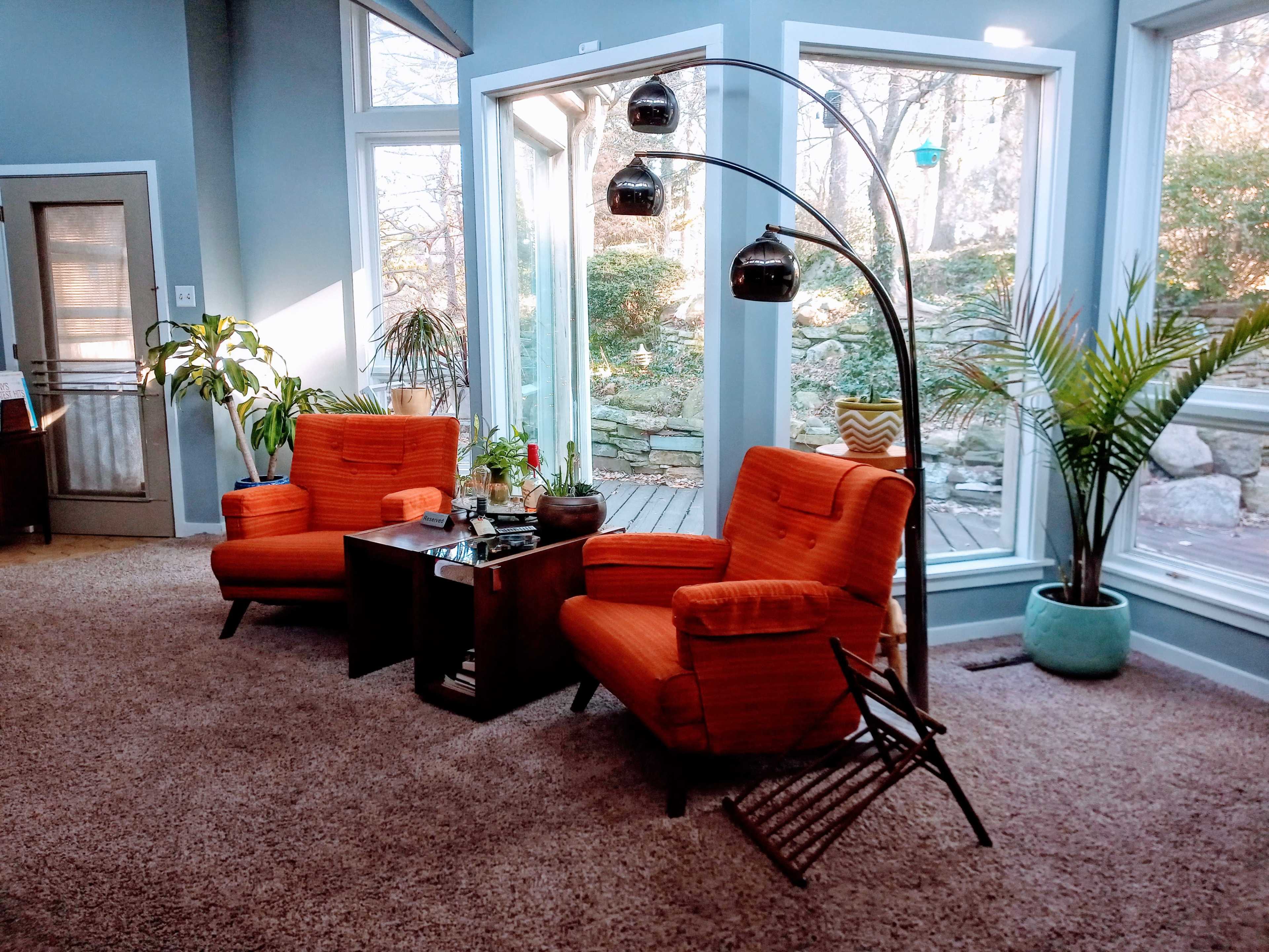Two orange armchairs positioned in a brightly lit room with large windows and a view of greenery outside.