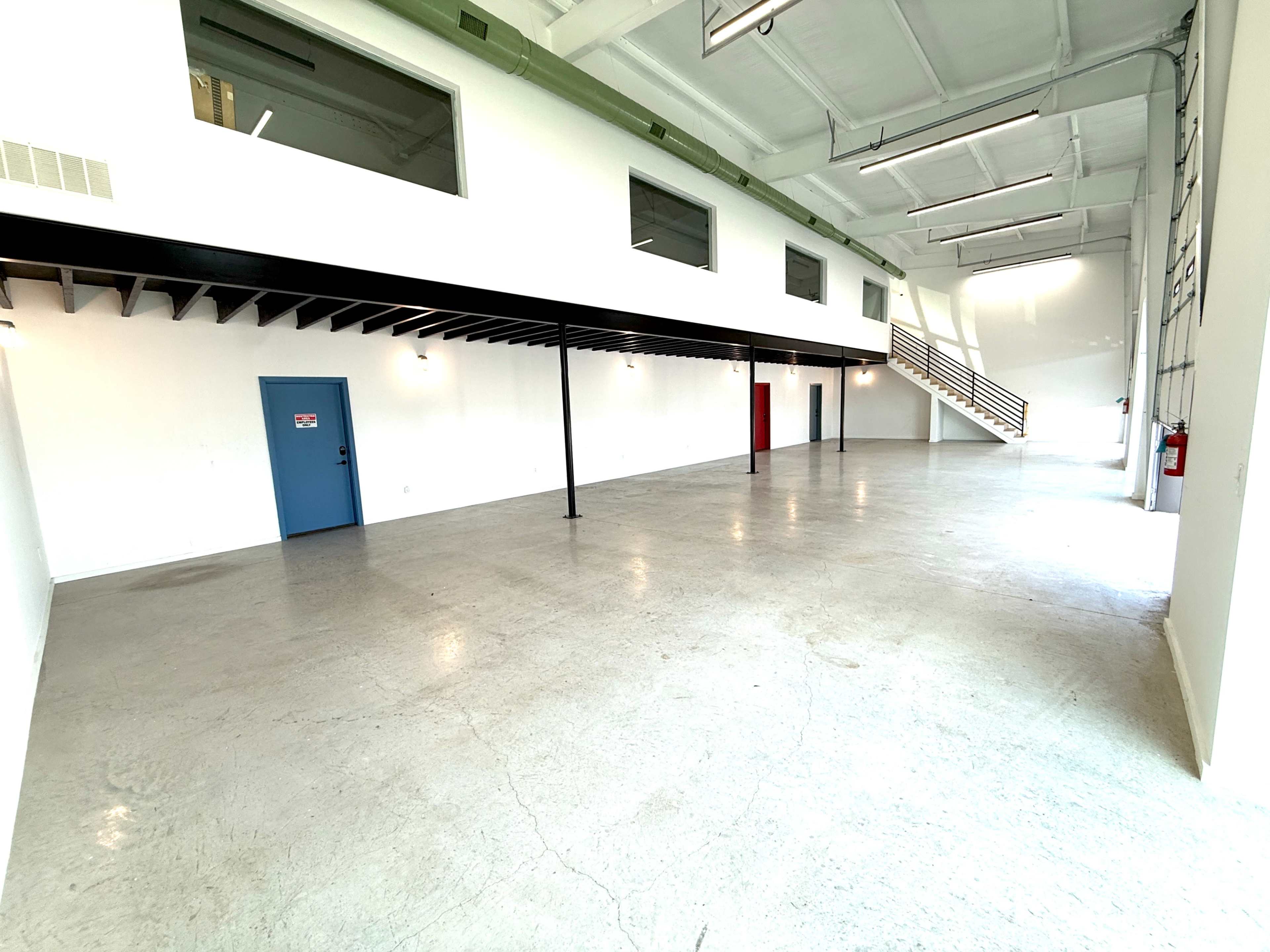 The image shows a spacious, empty room with polished concrete floors, white walls, and large windows, featuring a staircase at one end and a blue door on one side.
