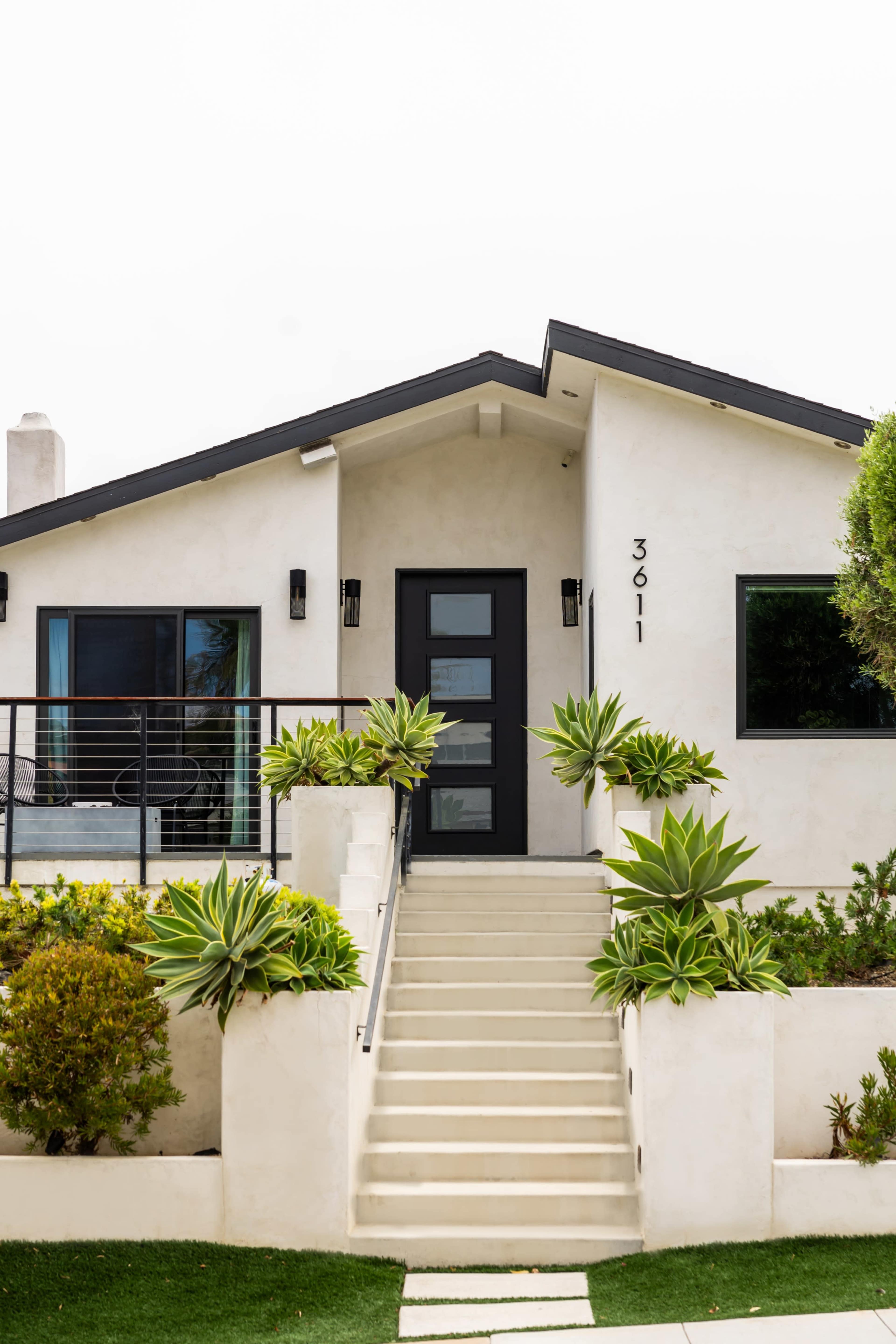 A modern house with a black door and potted plants flanks a set of stairs leading to the entrance.