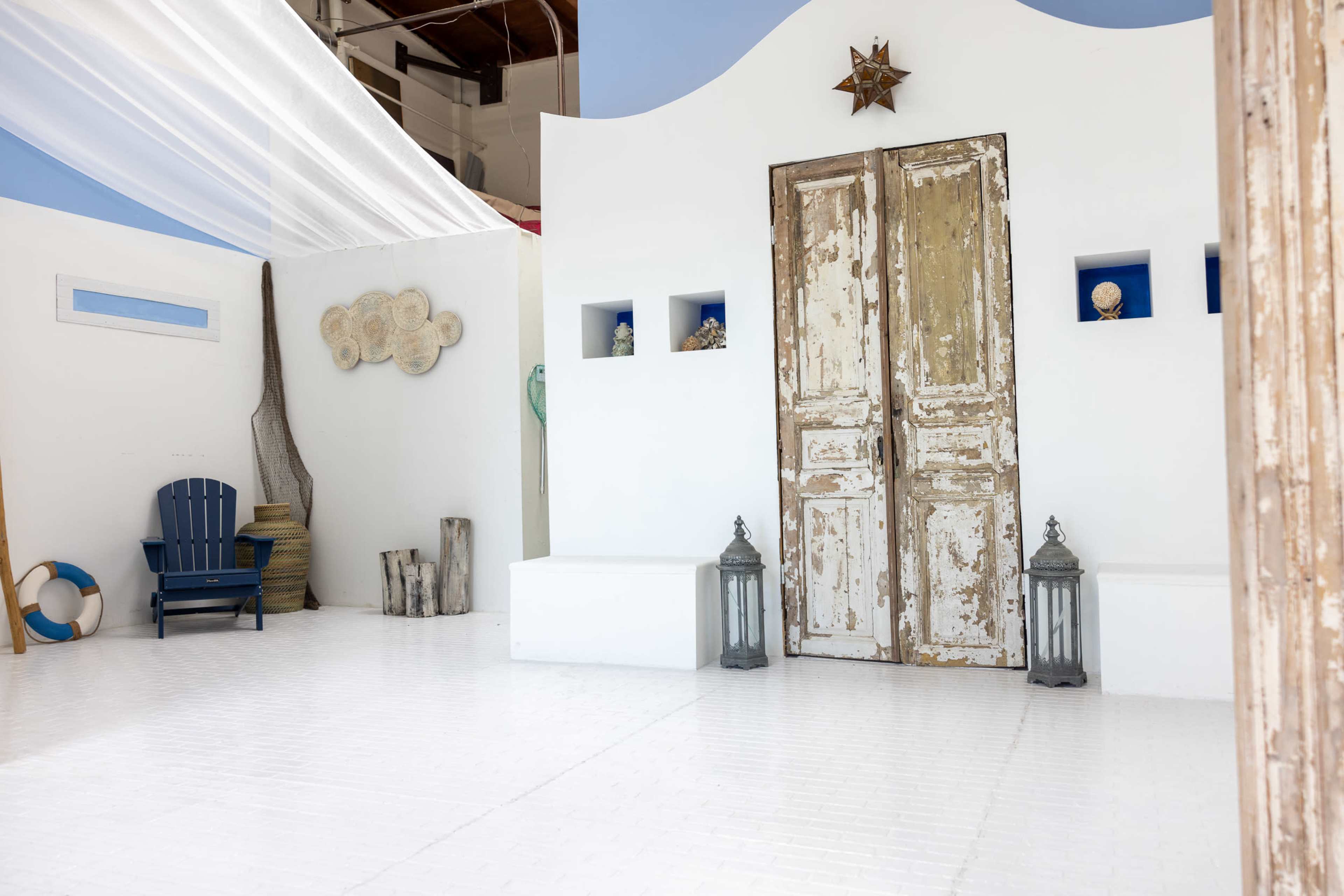 A minimalist indoor space features white walls, a pair of weathered double doors, and decorative elements such as lanterns and a wooden chair.