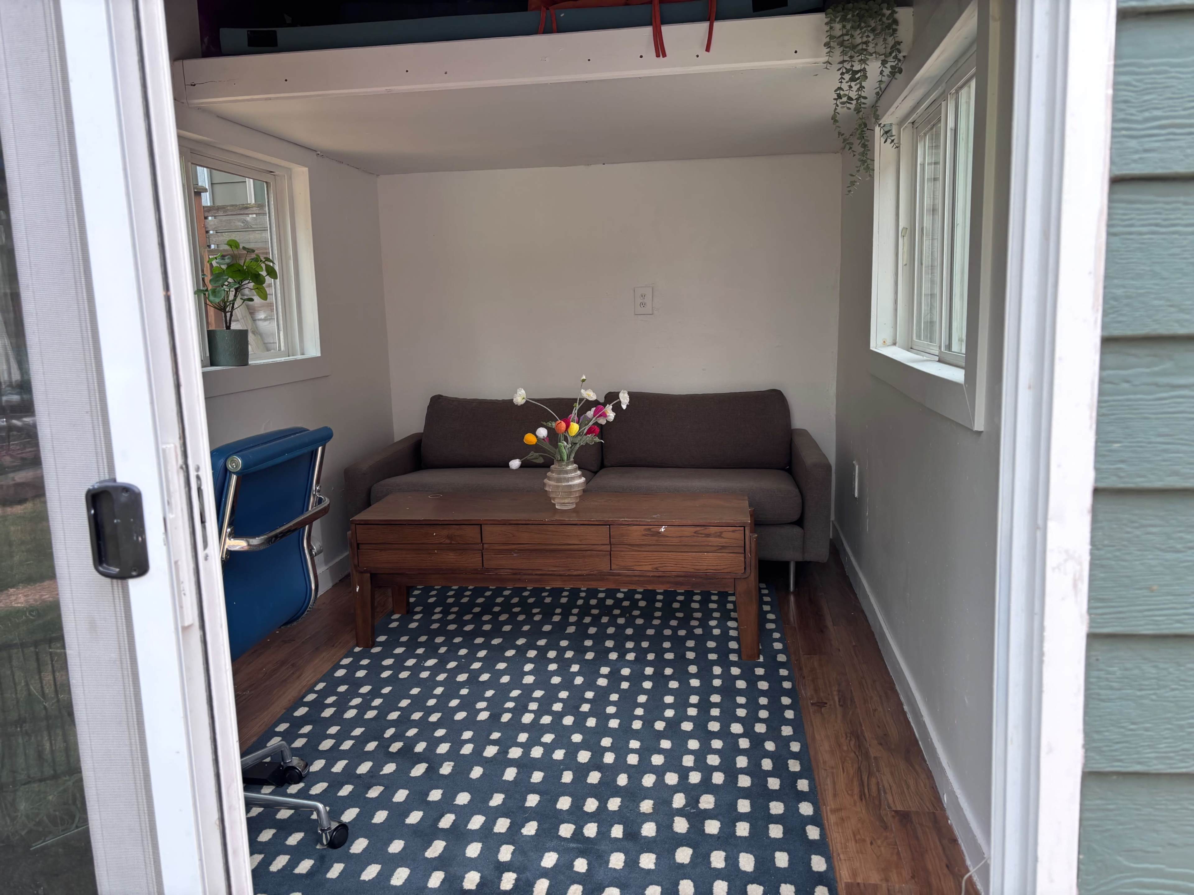 A small room features a gray sofa, a wooden coffee table with a vase of flowers, and a blue chair, all set against a patterned rug.