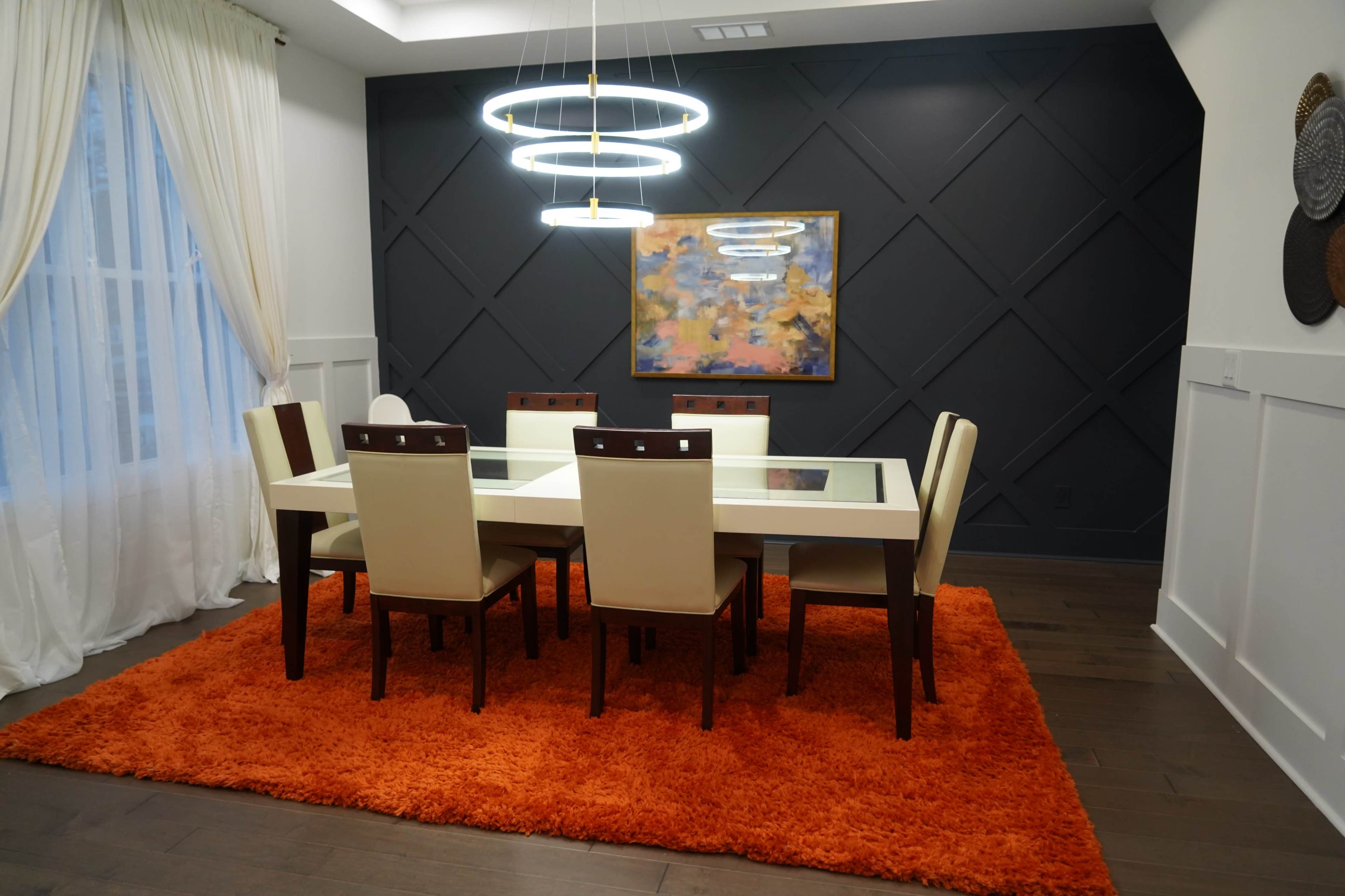 A modern dining room features a white table with glass top surrounded by light-colored chairs on an orange shag rug, with a geometric dark wall and a colorful artwork above.