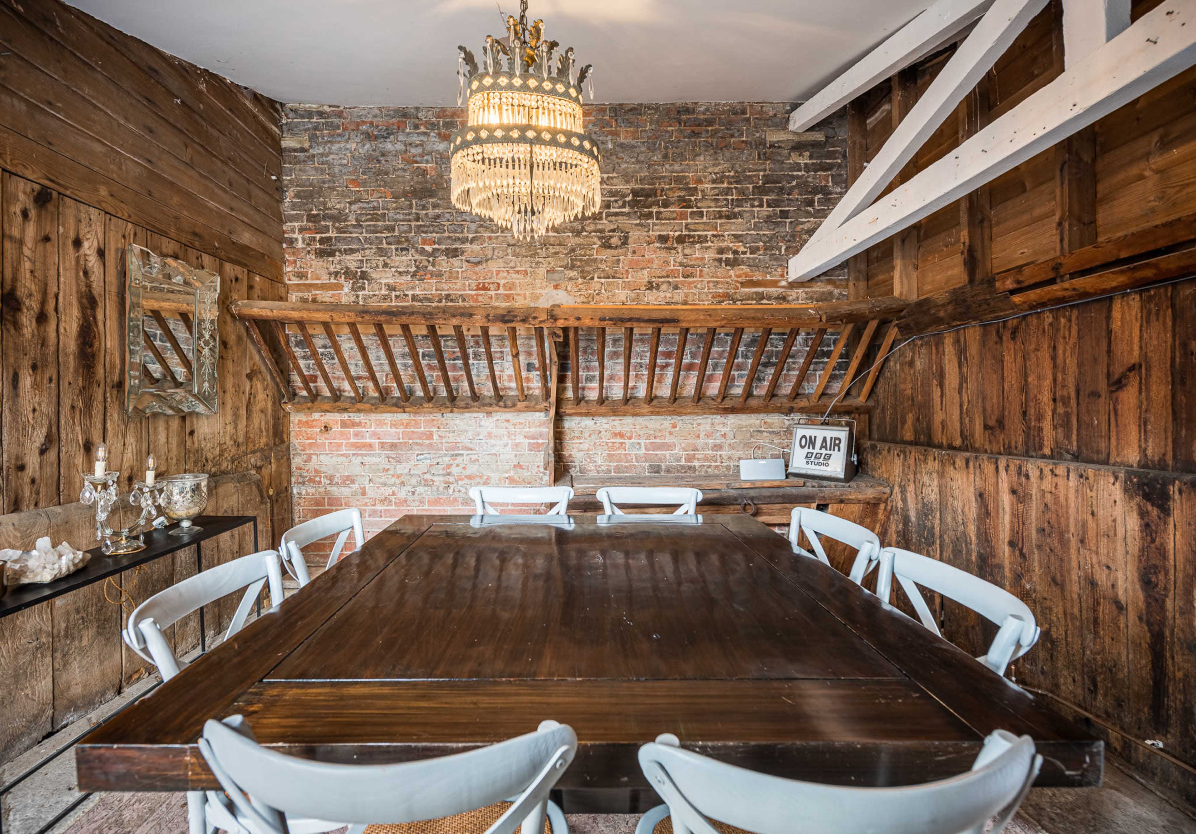 The image shows a rustic dining room featuring a large wooden table surrounded by eight chairs, with exposed brick walls and a chandelier hanging from the ceiling.