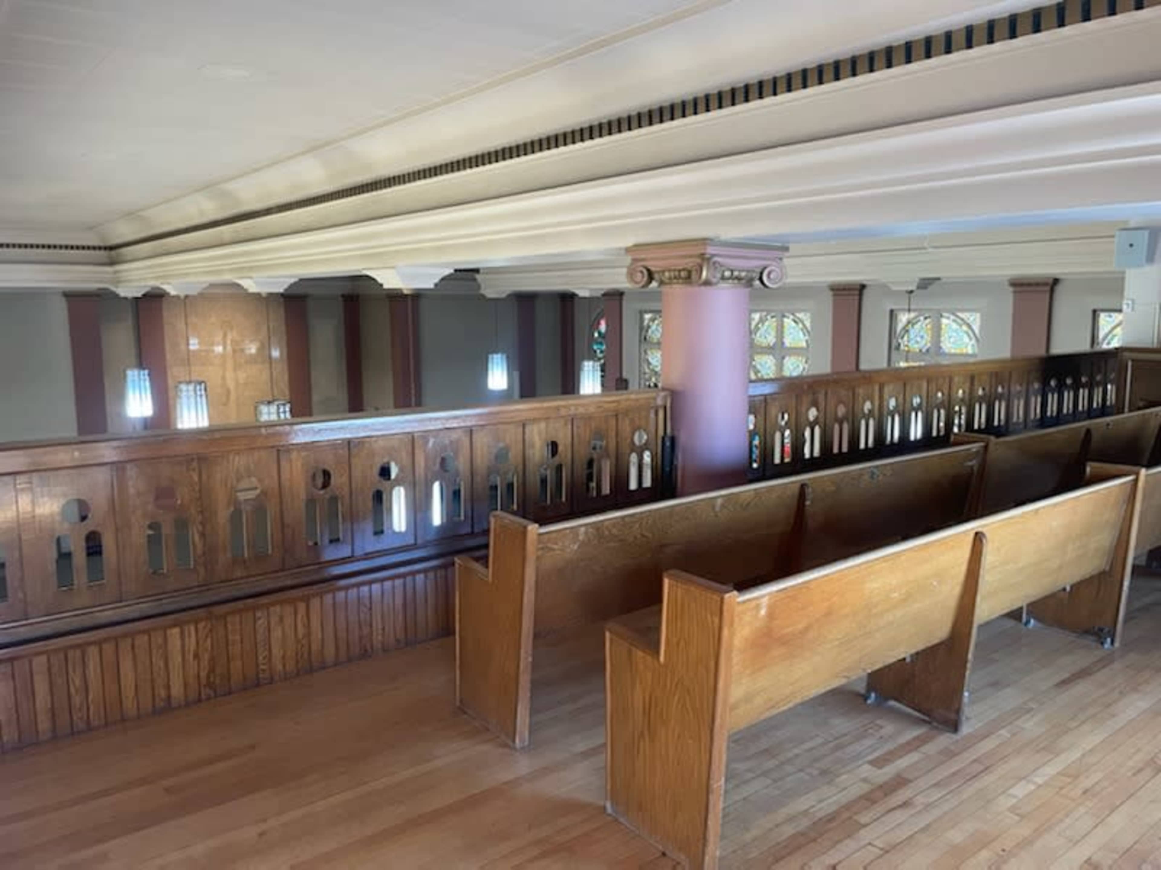 The image shows a wooden interior of a church balcony with several empty pews facing a railing and a set of stained glass windows in the background.