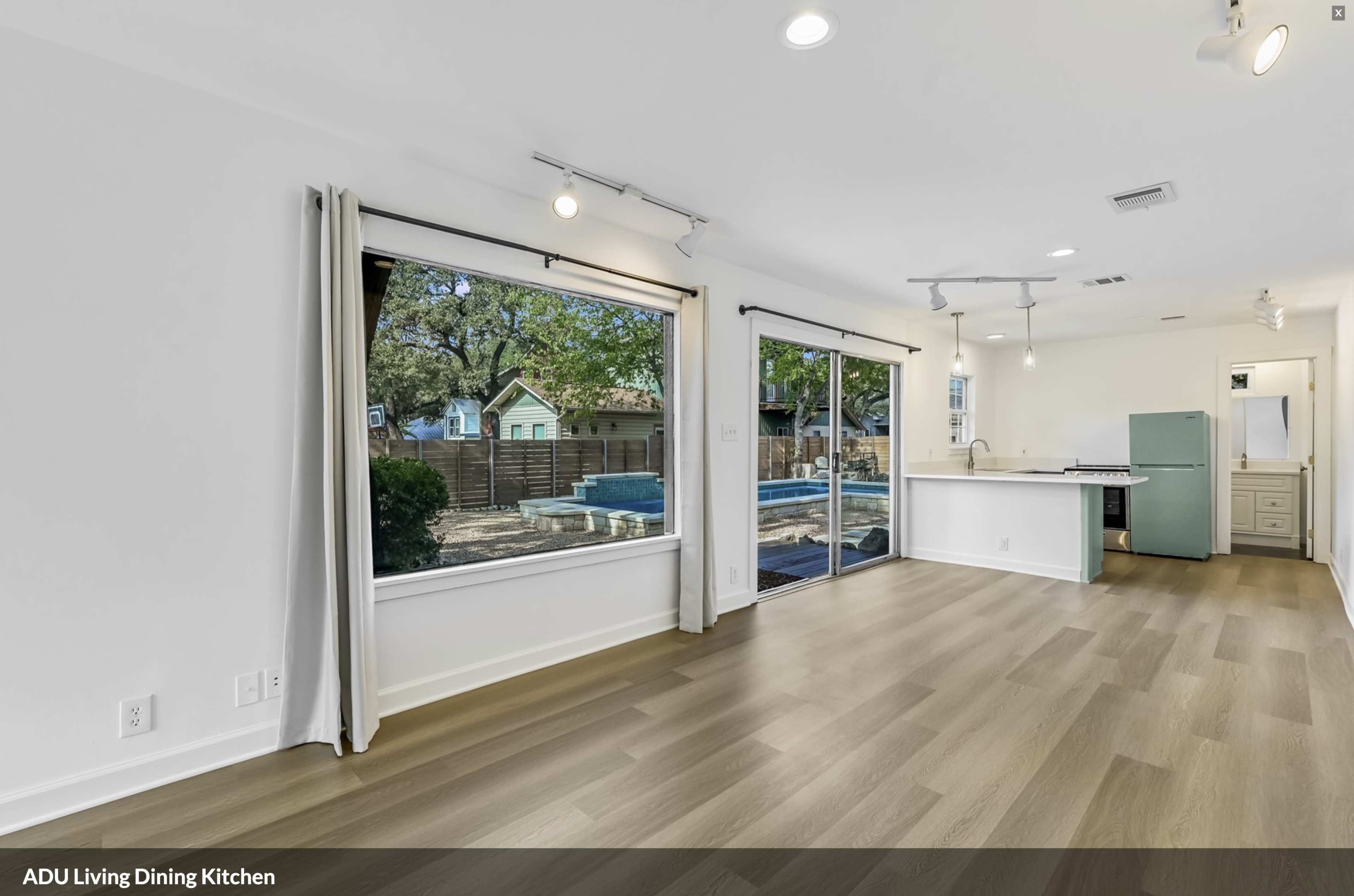 The image shows a modern open-concept living, dining, and kitchen area with large windows overlooking a backyard and a pool.