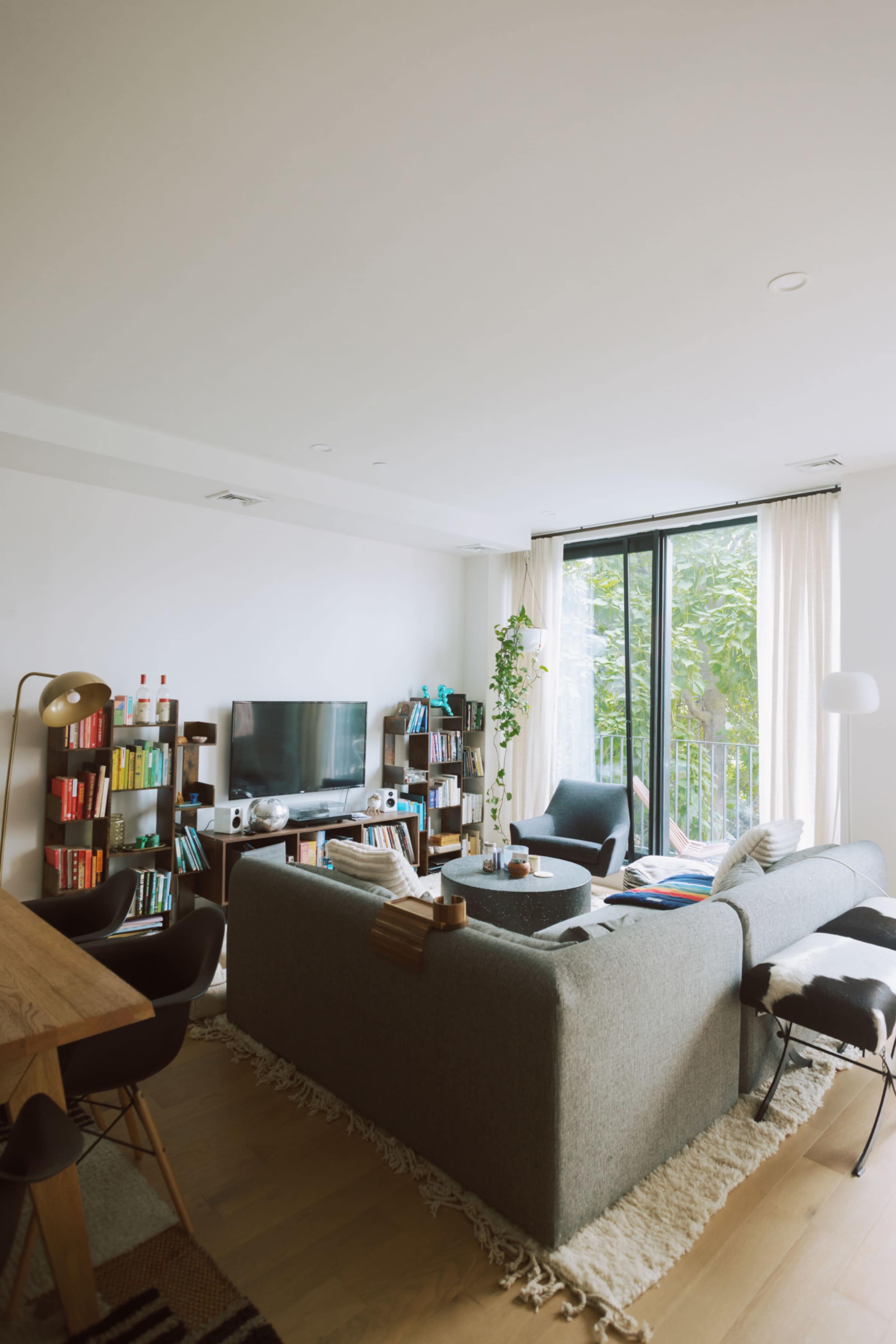 A modern living room features a gray sectional sofa, a wooden dining table, a TV on the wall, and shelves filled with books, while large windows let in natural light.