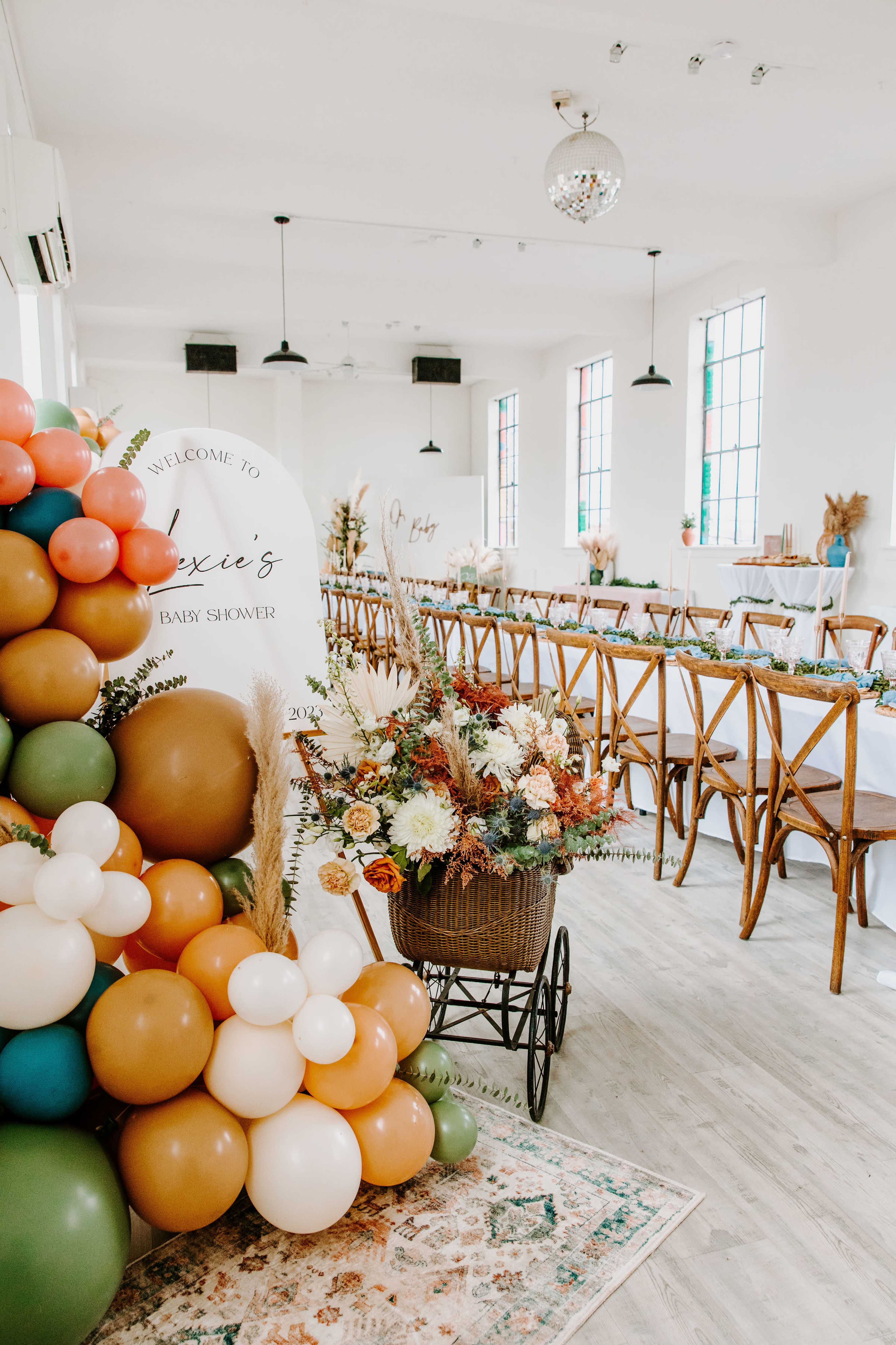 The image shows a beautifully decorated event space for a baby shower, featuring a long dining table set up with floral arrangements and a large display of balloons in various colors.