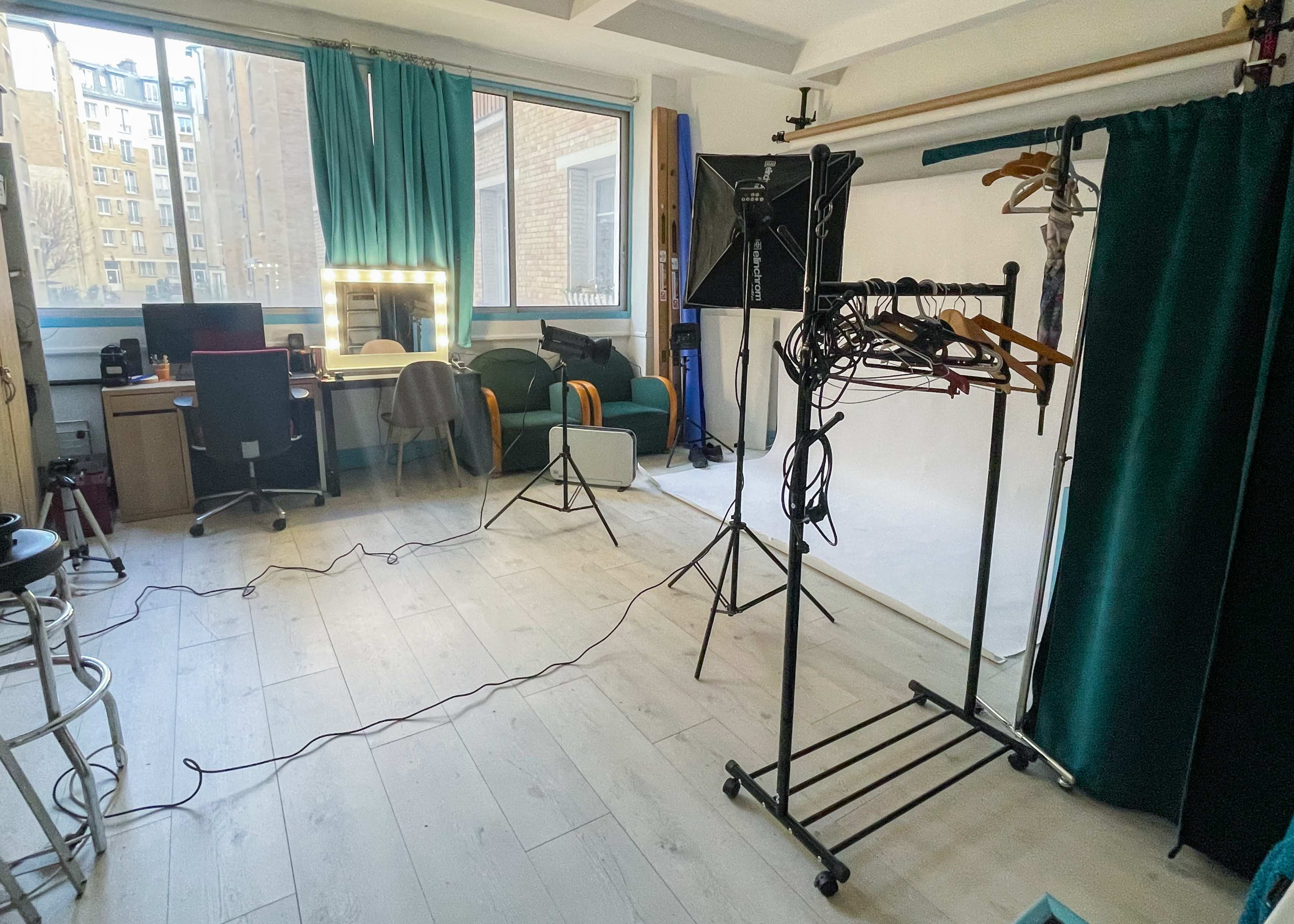 A well-lit studio space with a desk, a mirror, a green chair, a clothing rack, and a backdrop setup.