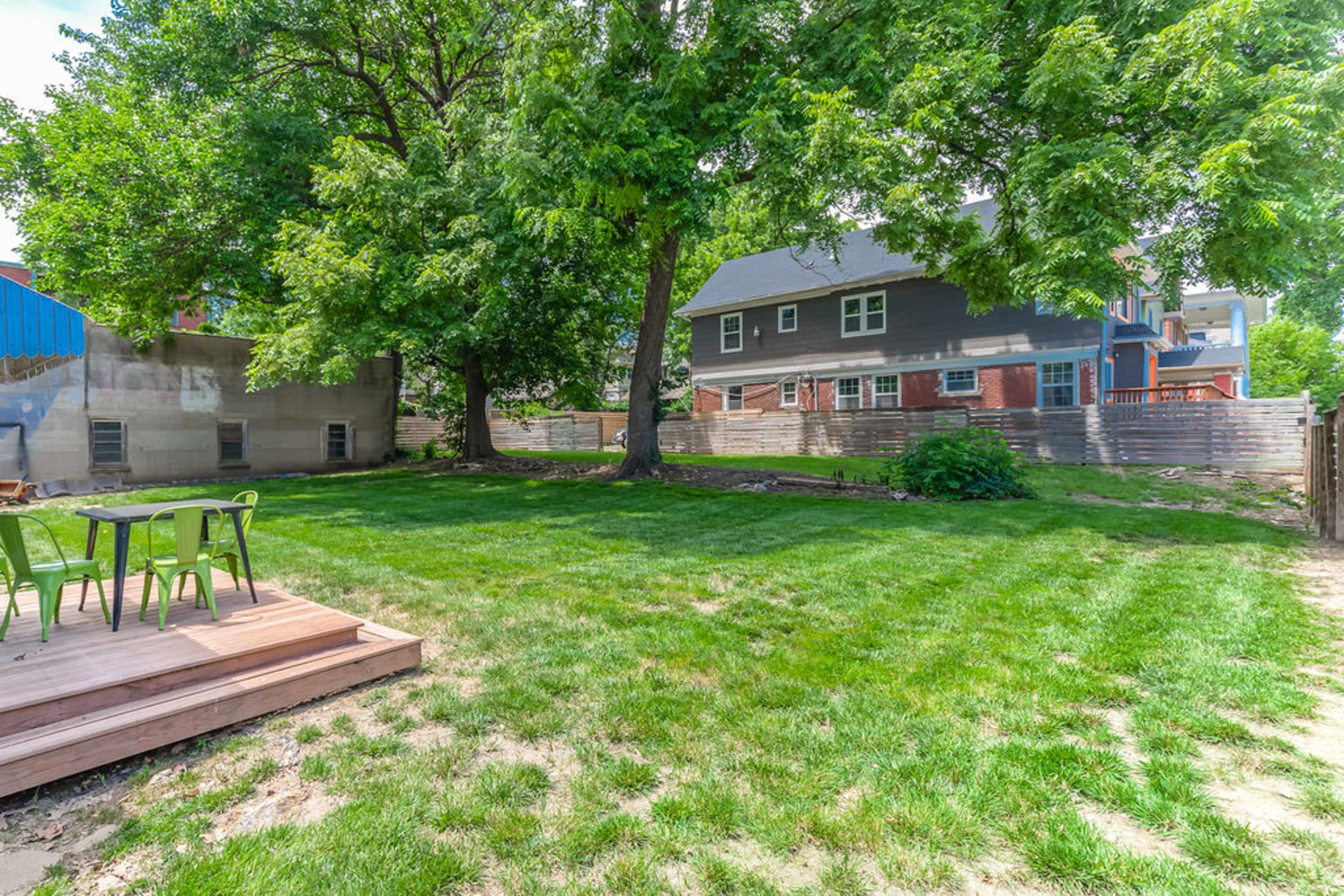 A spacious backyard with a wooden deck, green grass, and several large trees, alongside a two-story house in the background.