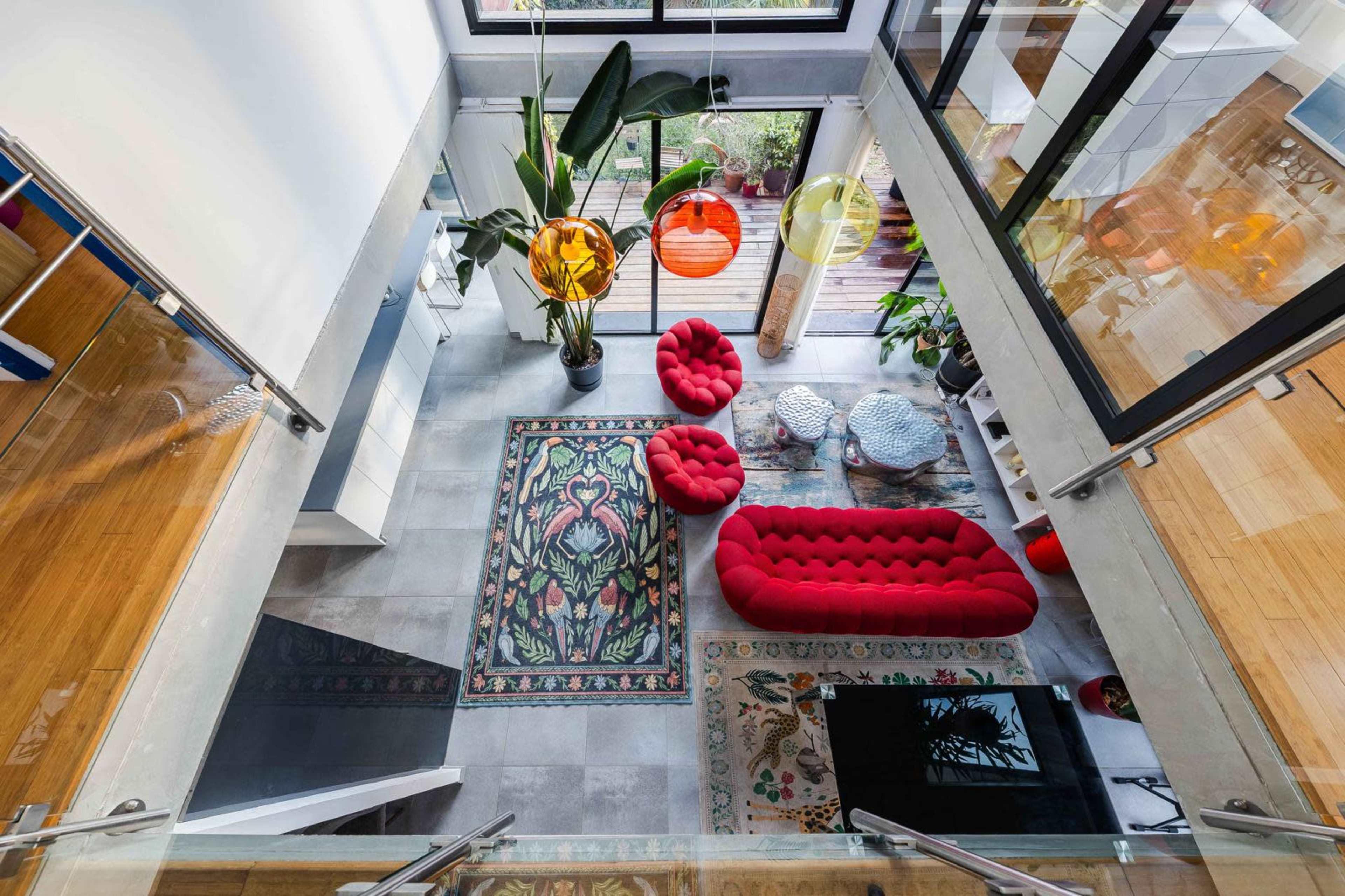 The image shows a modern living room from an overhead perspective, featuring a red tufted sofa, colorful pendant lights, and various plants arranged around patterned rugs.