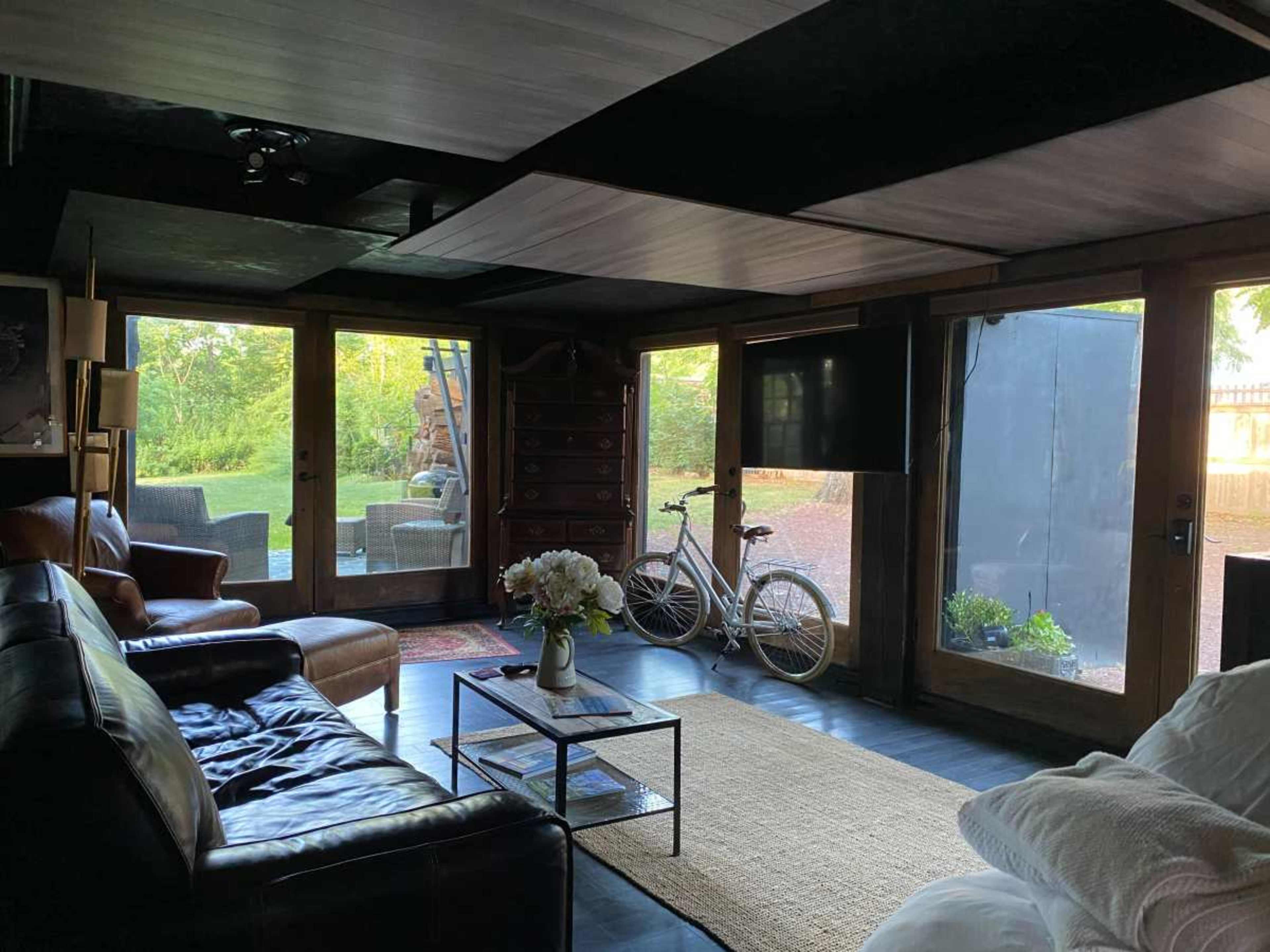 The image shows a modern living room with dark wooden beams, a sectional couch, a coffee table, a vintage bicycle, and large glass doors opening to an outdoor area.