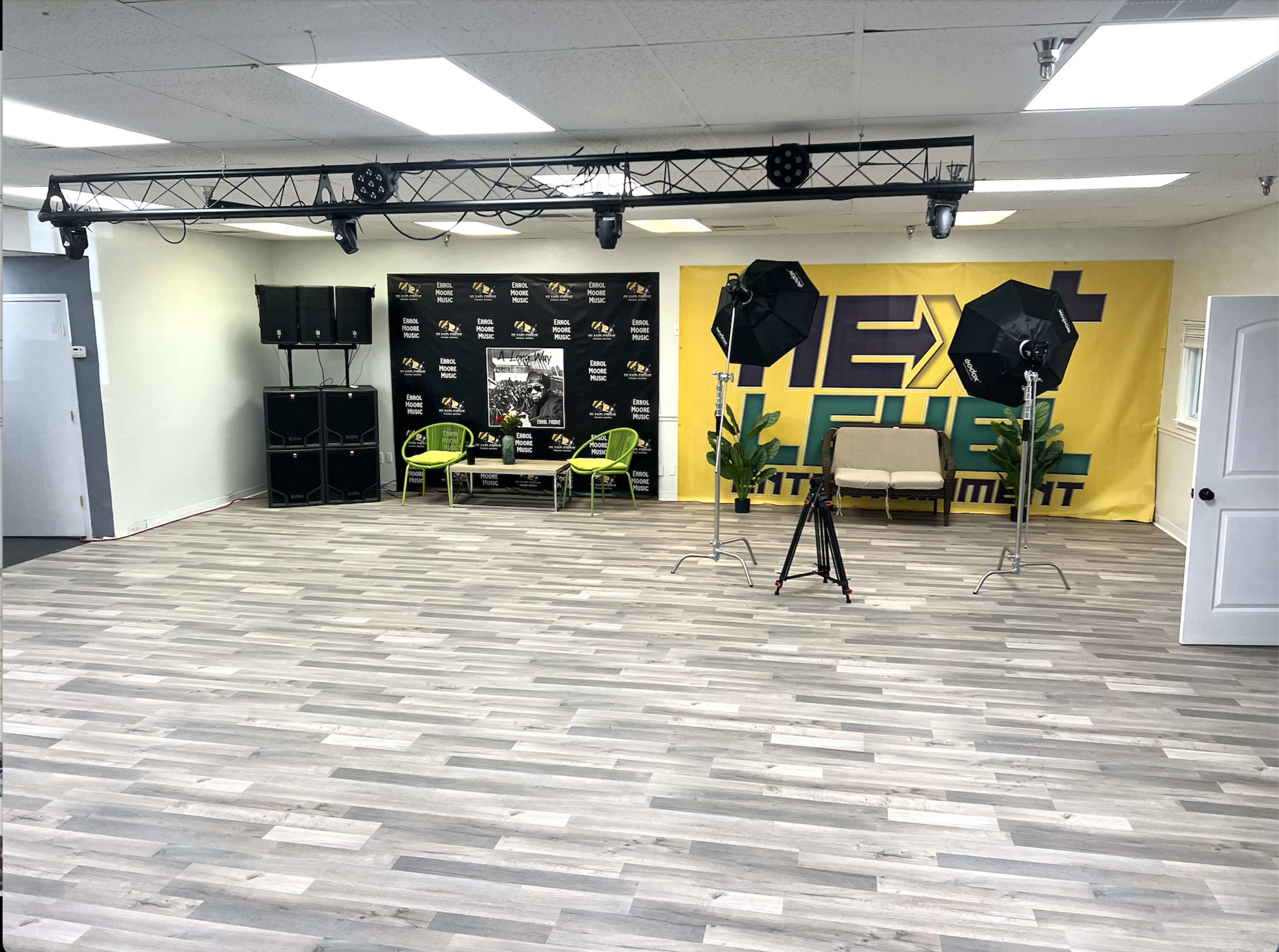 An empty room with wooden flooring, staged for a presentation, featuring a large yellow banner, two green chairs, and studio lighting equipment.