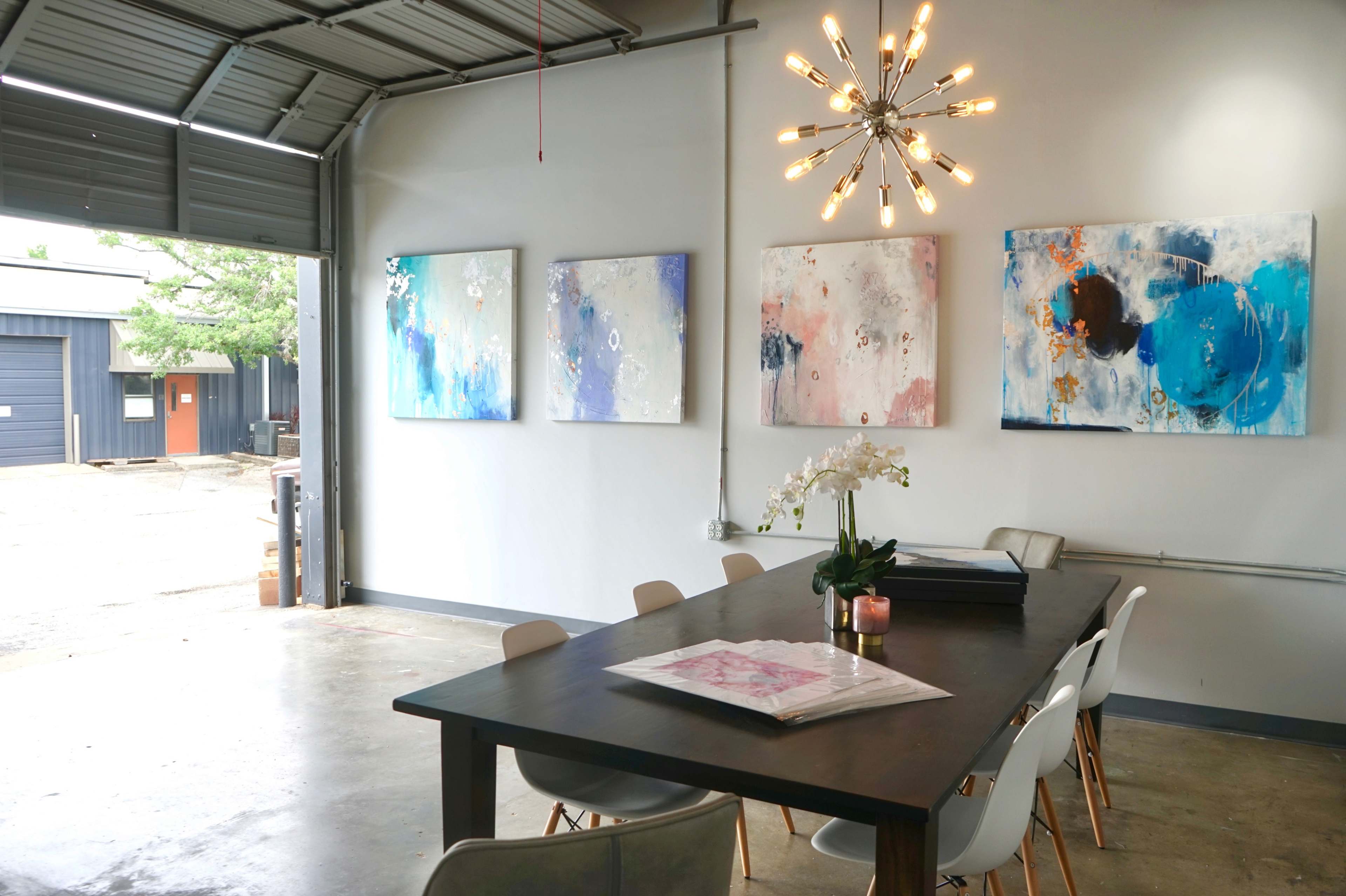 A modern meeting room with a large table, surrounded by chairs, and four colorful abstract paintings on the wall, illuminated by a unique light fixture.