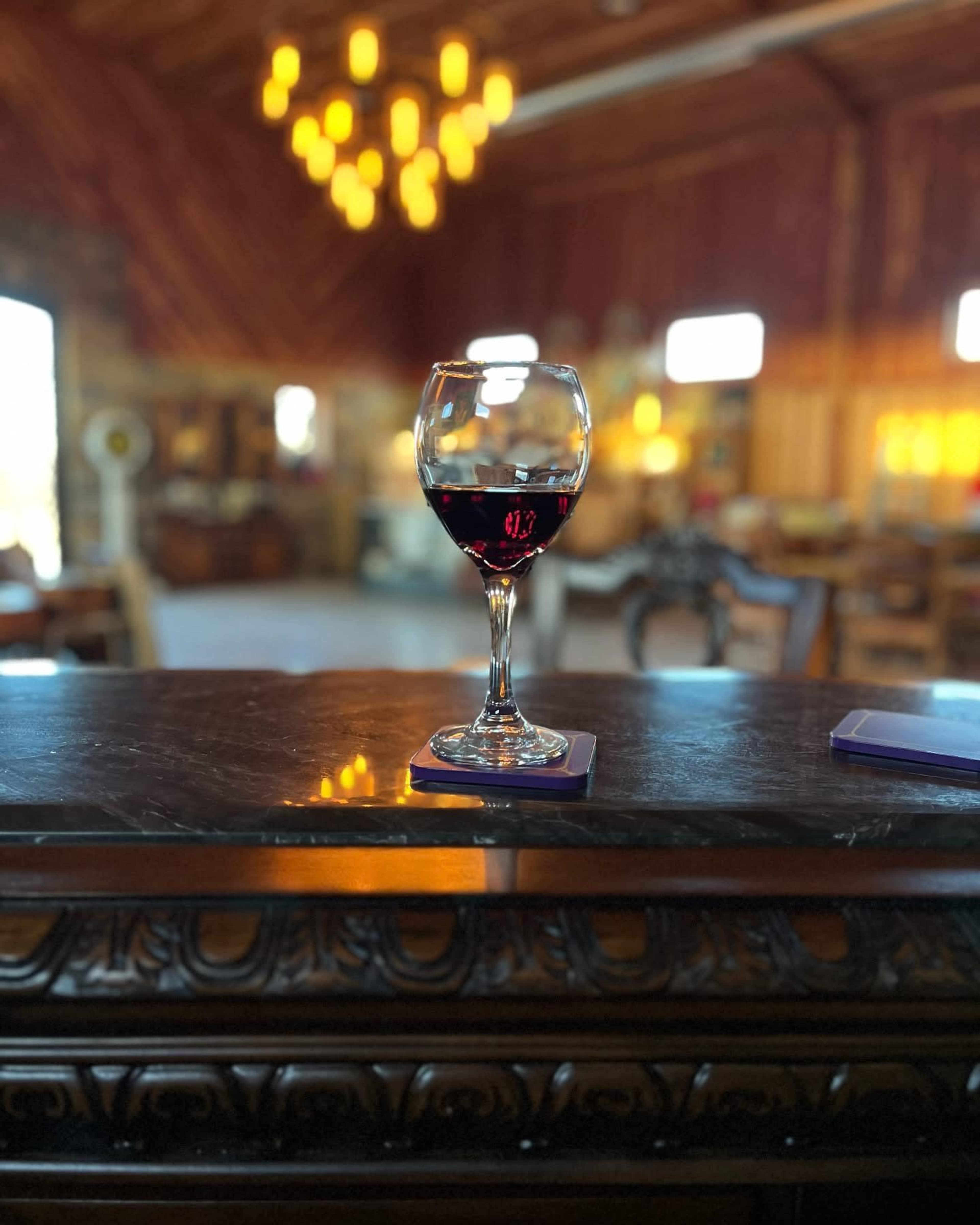 A glass of red wine sits on a coaster atop a dark wooden bar, illuminated by warm lights in a rustic interior.