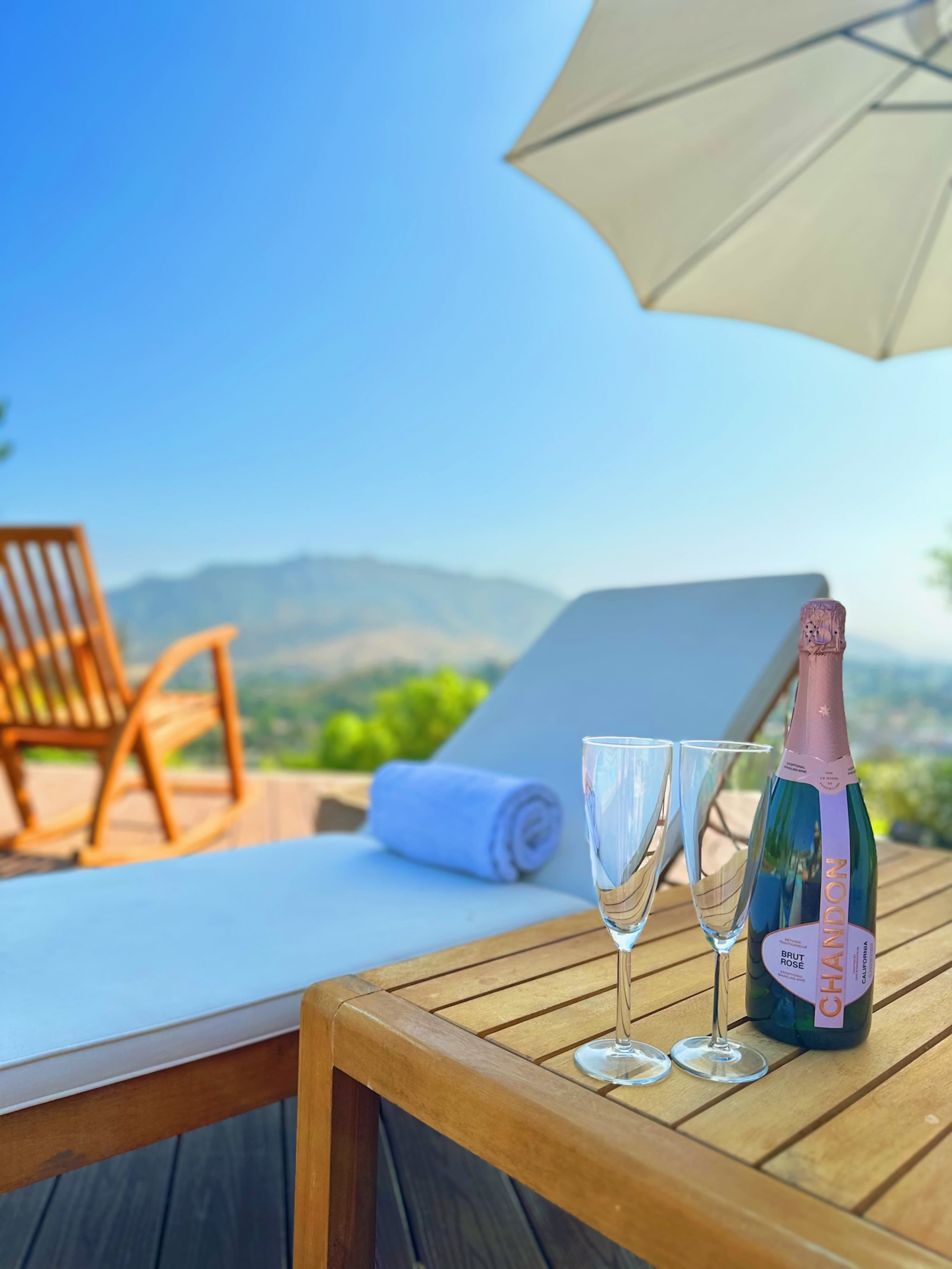 A bottle of champagne and two glasses rest on a wooden table beside a lounge chair and a towel under an umbrella with a view of mountains in the background.