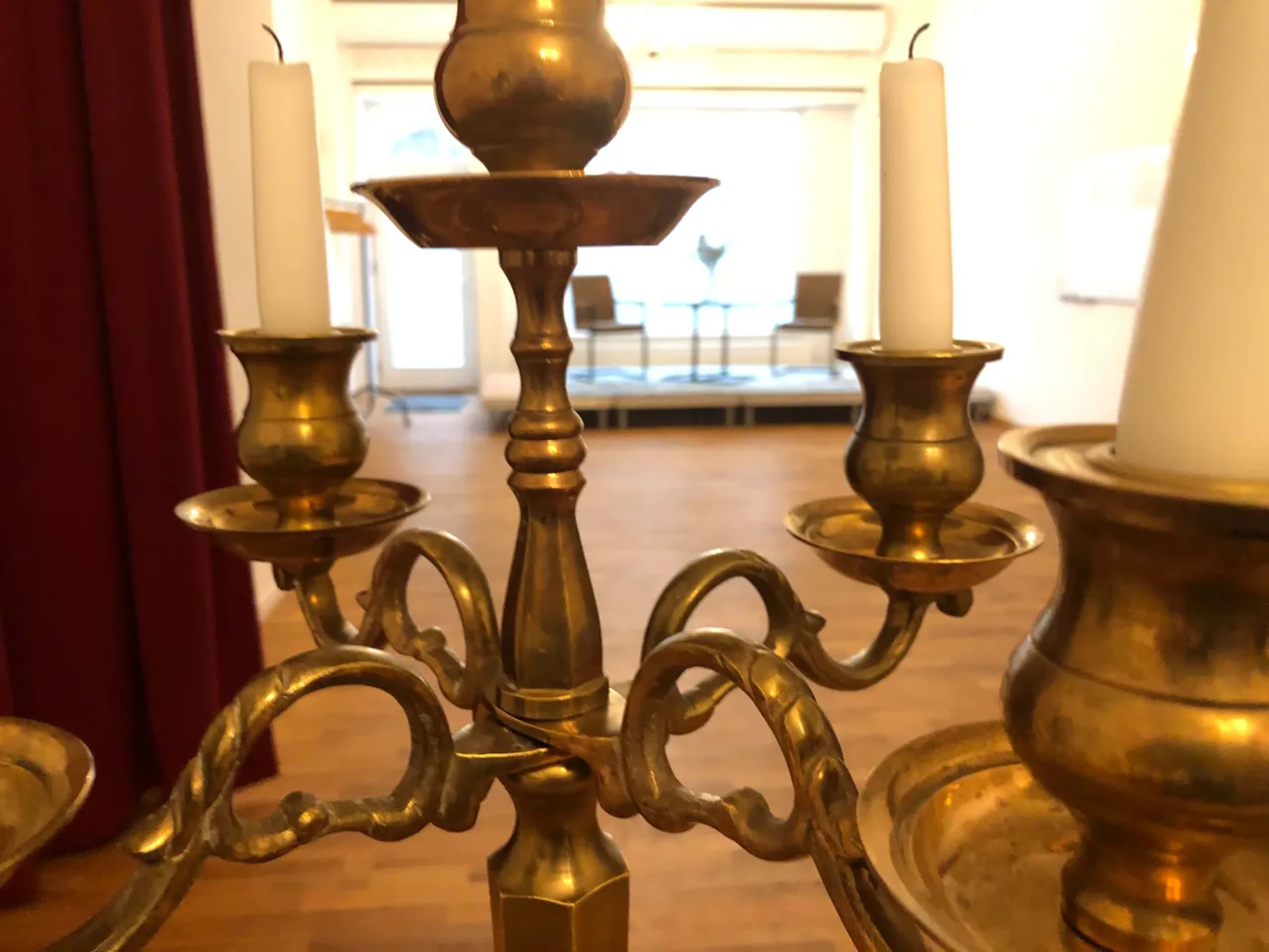 The image shows a brass chandelier with four candle holders and white candles, positioned in a room with wooden flooring and distant seating.