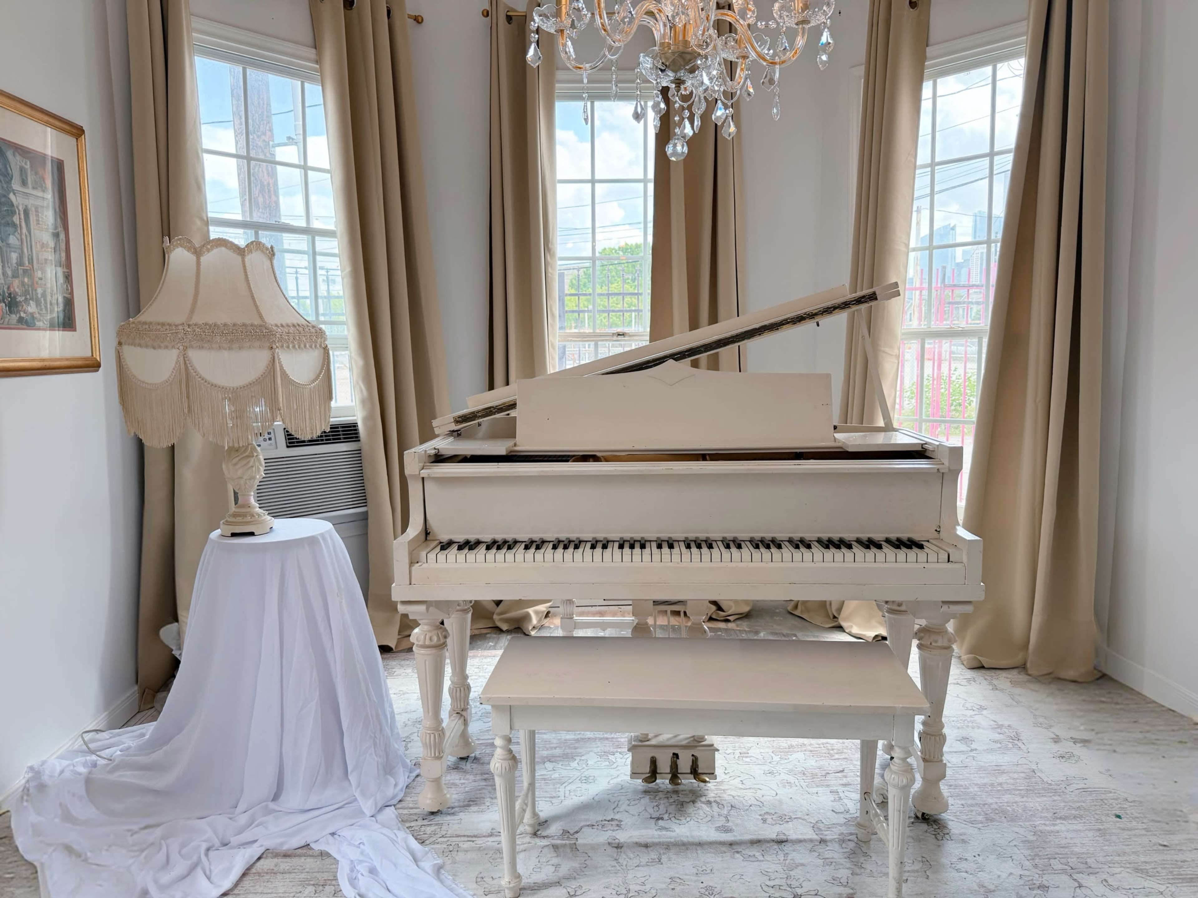 A white piano with its lid propped open is positioned next to a lamp on a draped table in a bright room with large windows and curtains.
