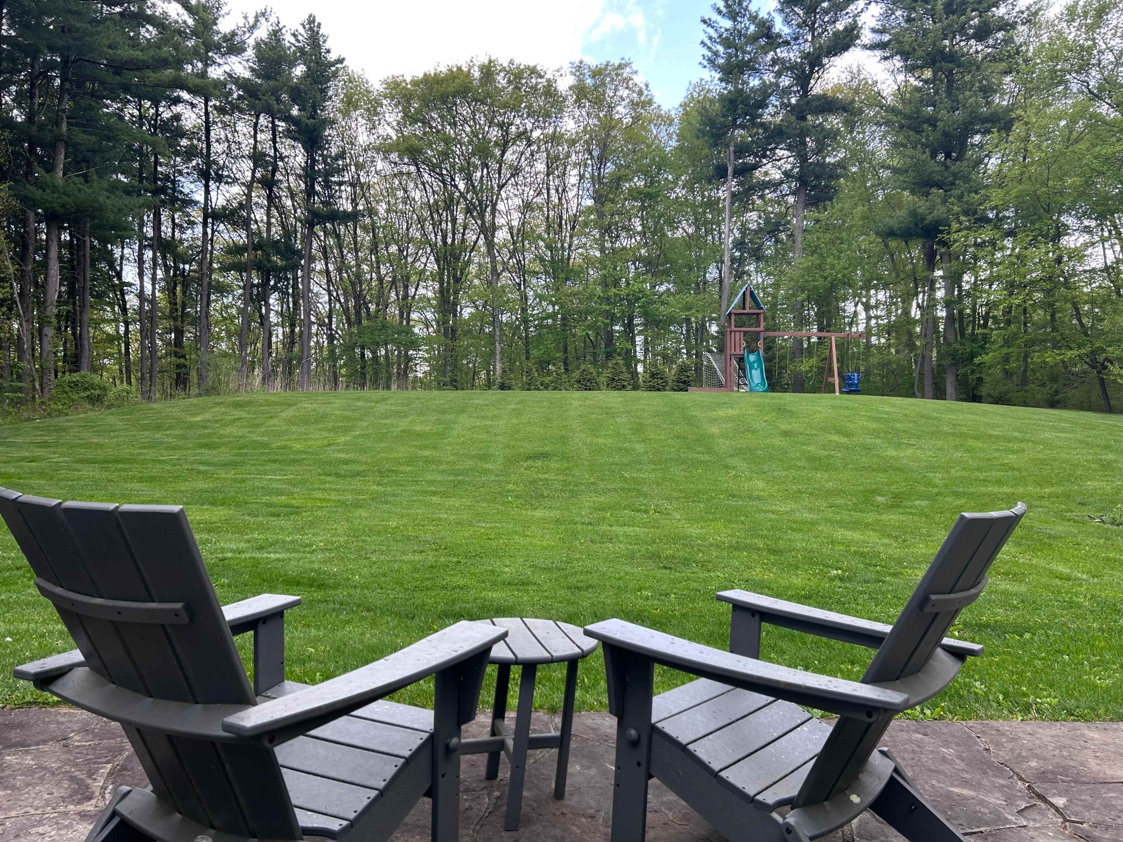 The image shows two black adirondack chairs and a small table on a patio, overlooking a neatly mowed green lawn with a playground structure in the background and trees lining the horizon.