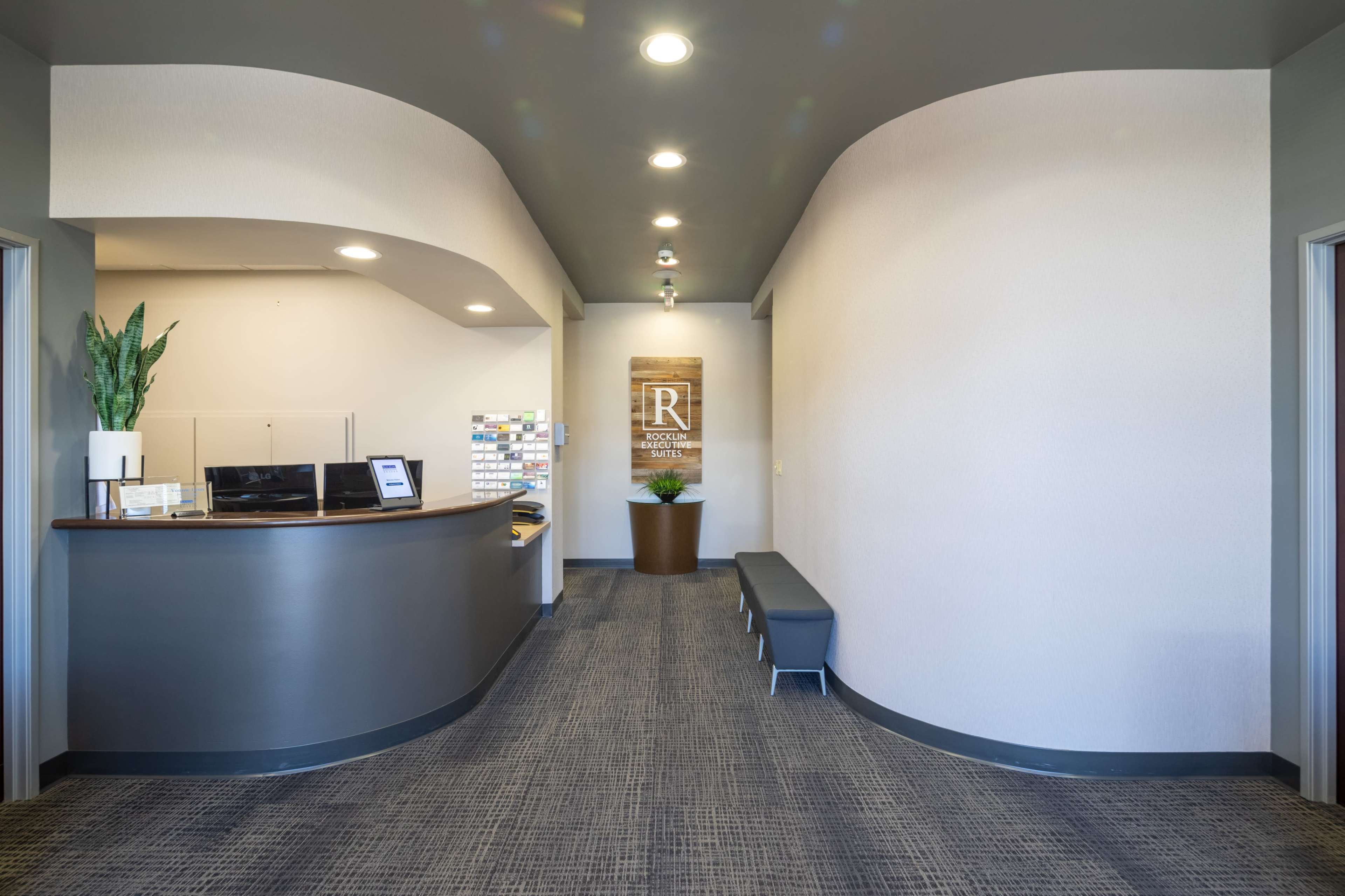 The image shows a modern reception area featuring a curved front desk, a seating bench, and a potted plant along a well-lit corridor.