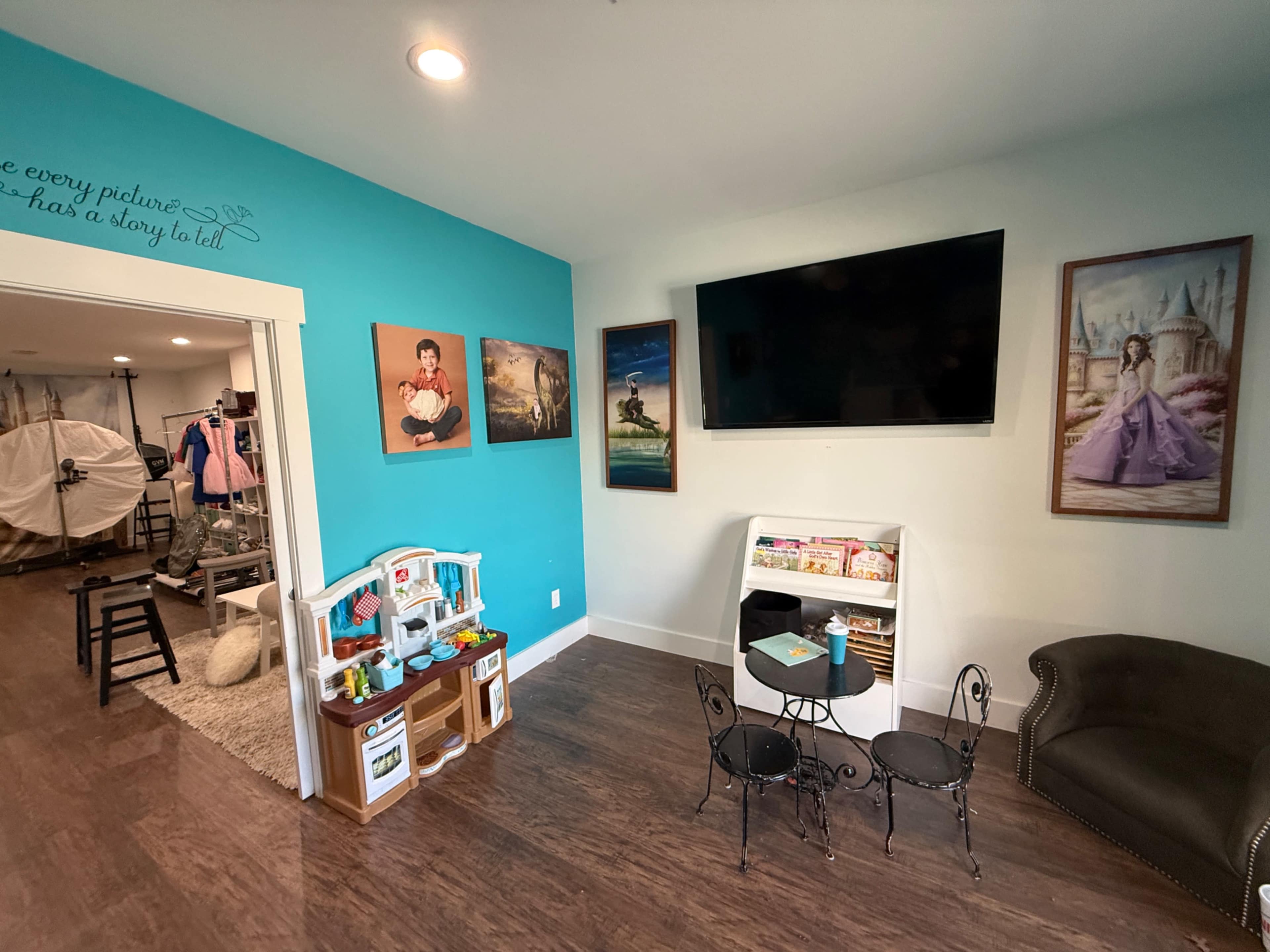 The image shows a brightly colored room with a turquoise wall, featuring children's toys, a small table and chairs, a television, and artwork of people on the walls.