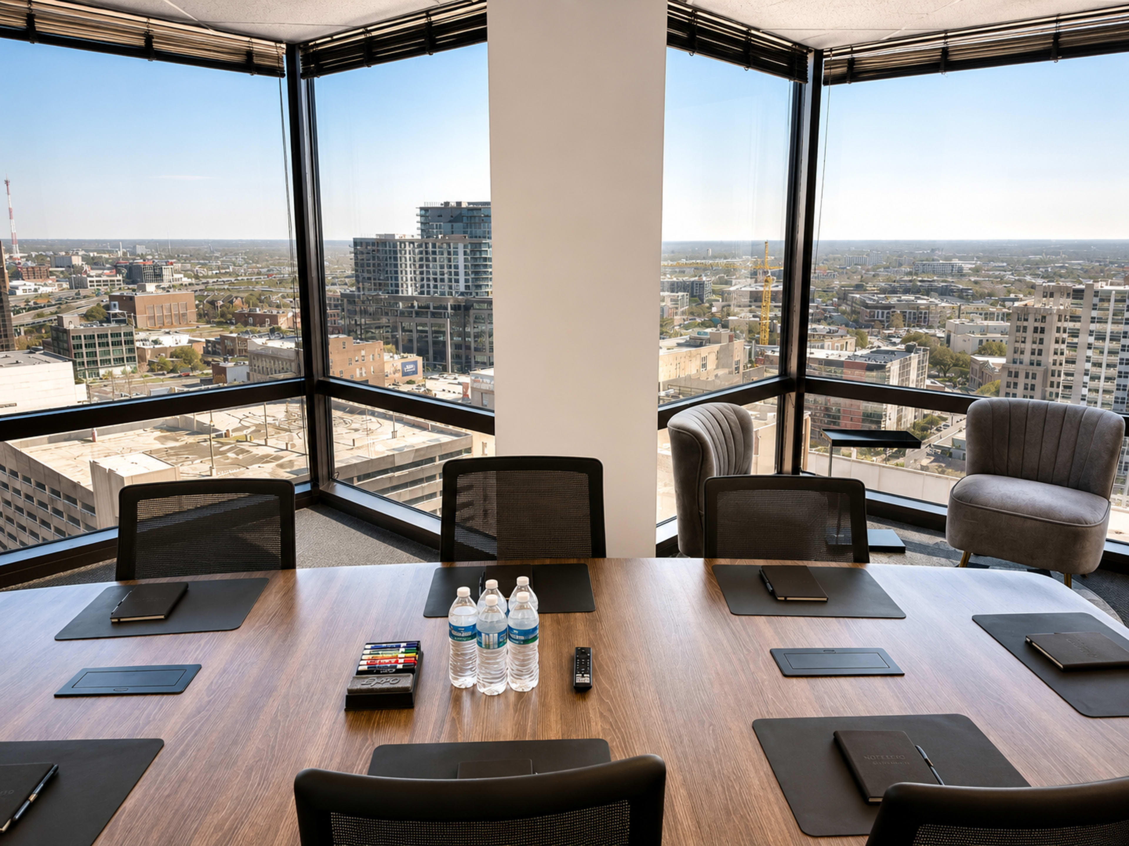 A large conference room featuring a wooden table surrounded by black chairs, with water bottles and notepads on the table, and floor-to-ceiling windows offering a view of the city skyline.