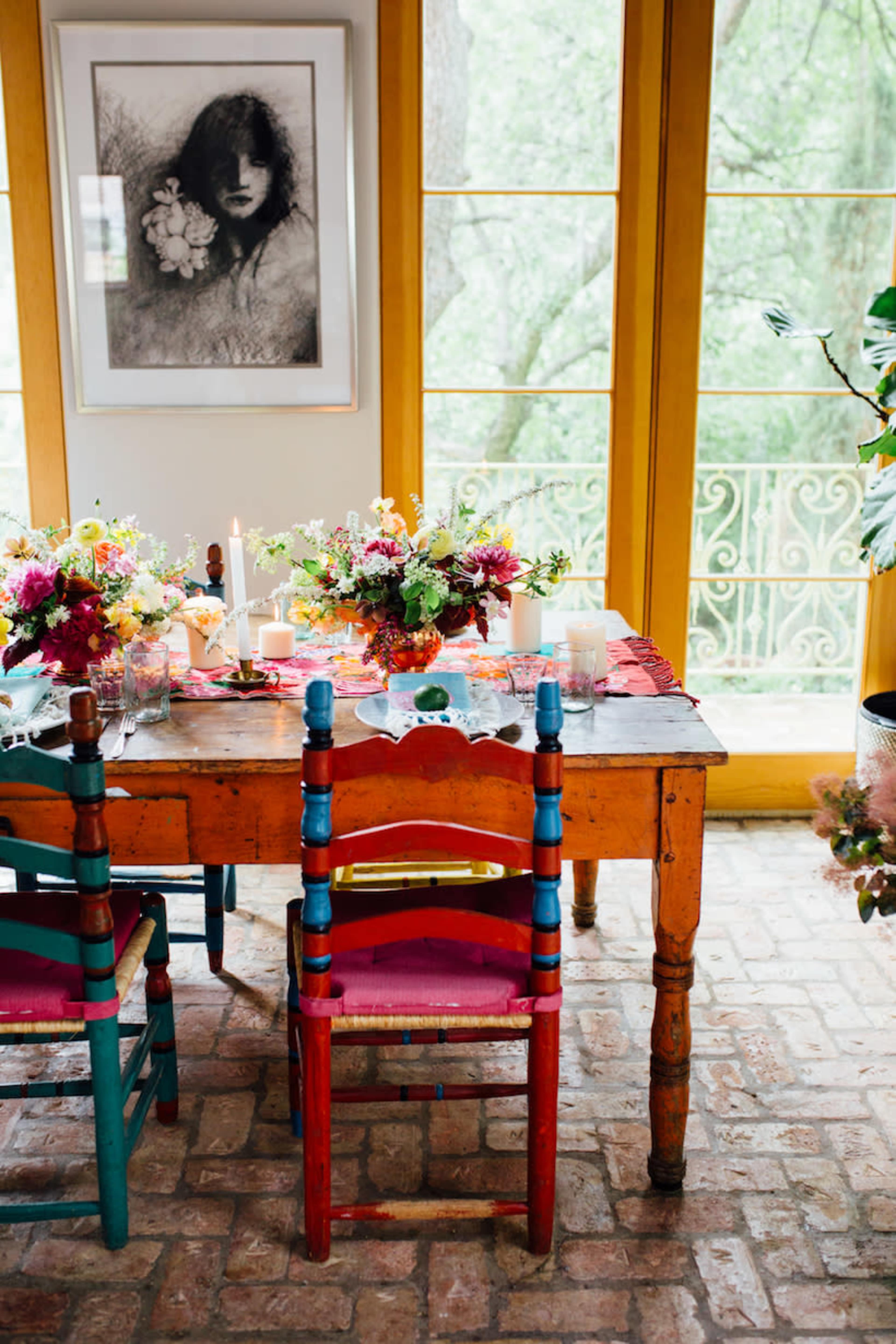 A colorful wooden dining table is set with floral arrangements and candles, surrounded by uniquely painted chairs, in a bright room with large windows overlooking greenery.
